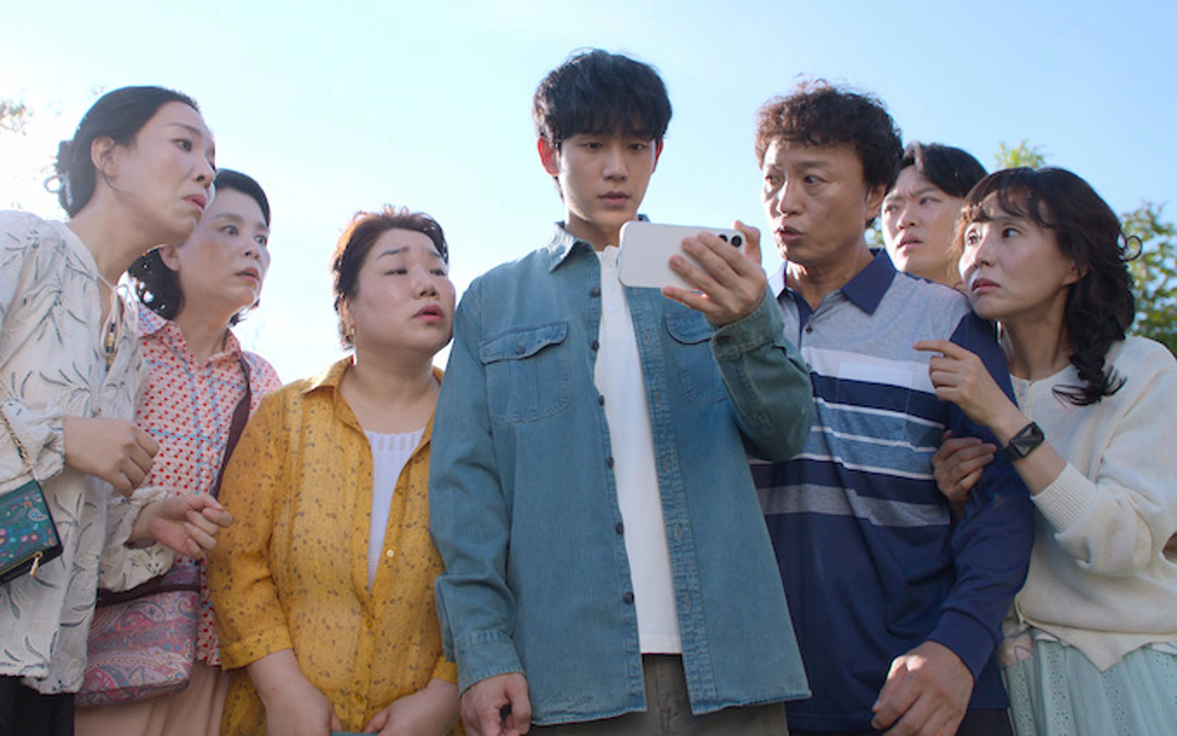 Jung Hae-in looks at a phone while a group of villagers crowds around him outdoors.