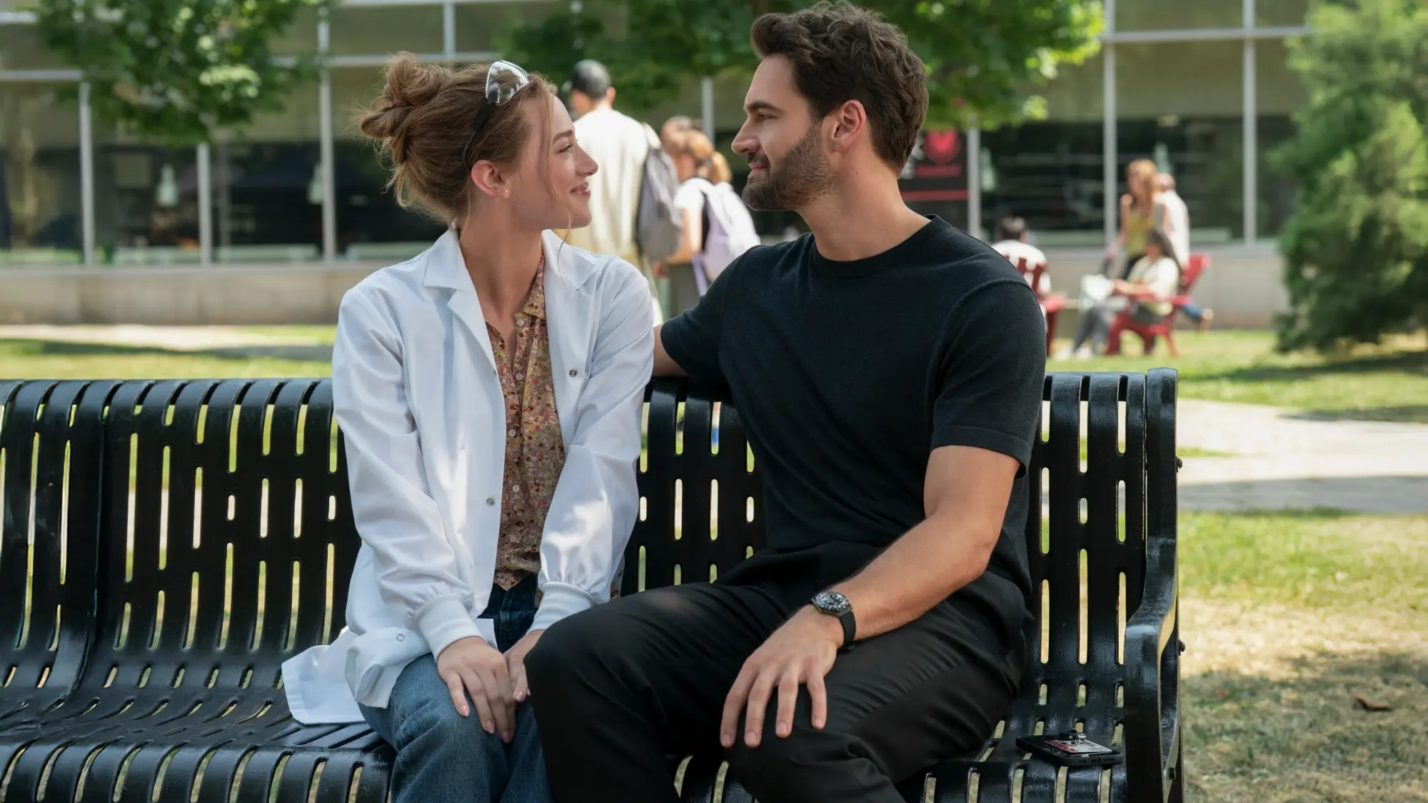 Lili Reinhart and Tom Bateman look at each other on a park bench in The Love Hypothesis.