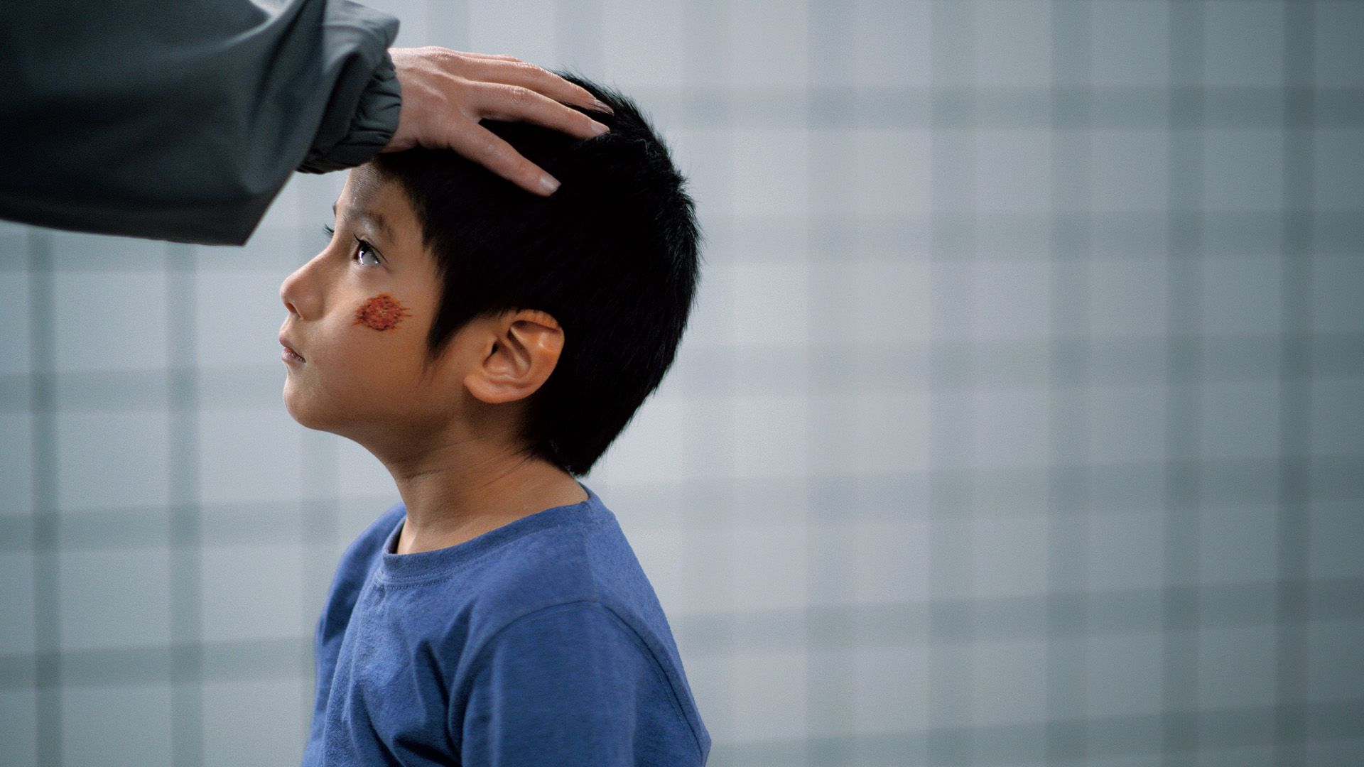 A boy in a blue shirt looks up as an adult rests a hand on his head.