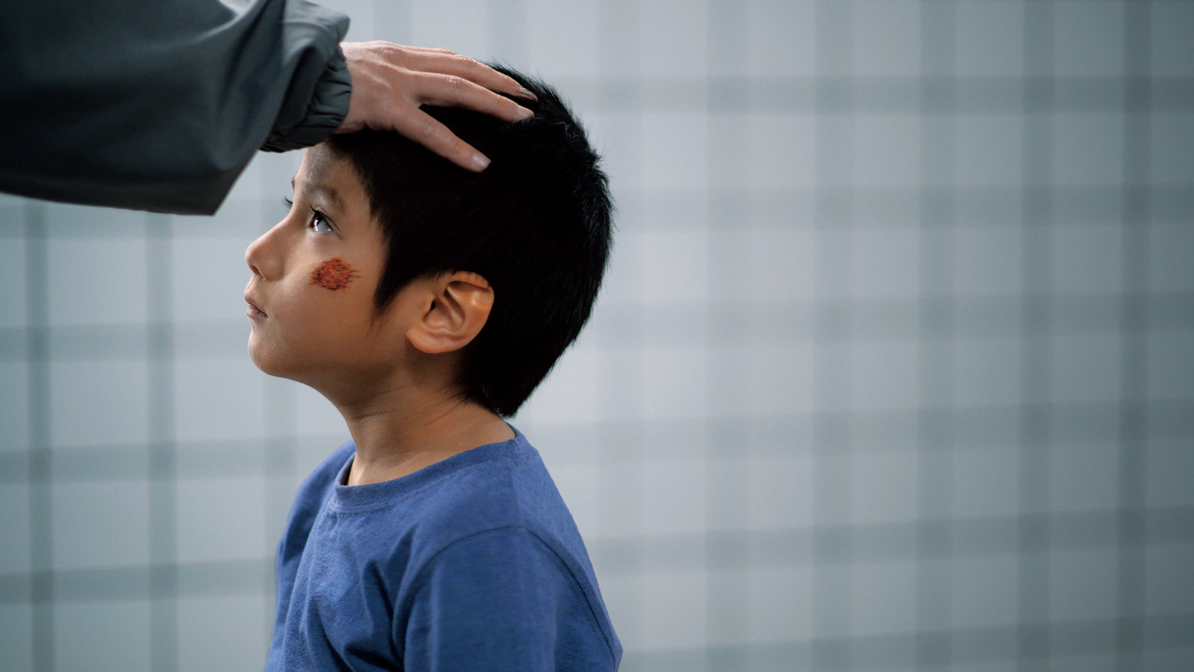 A boy in a blue shirt looks up as an adult rests a hand on his head.