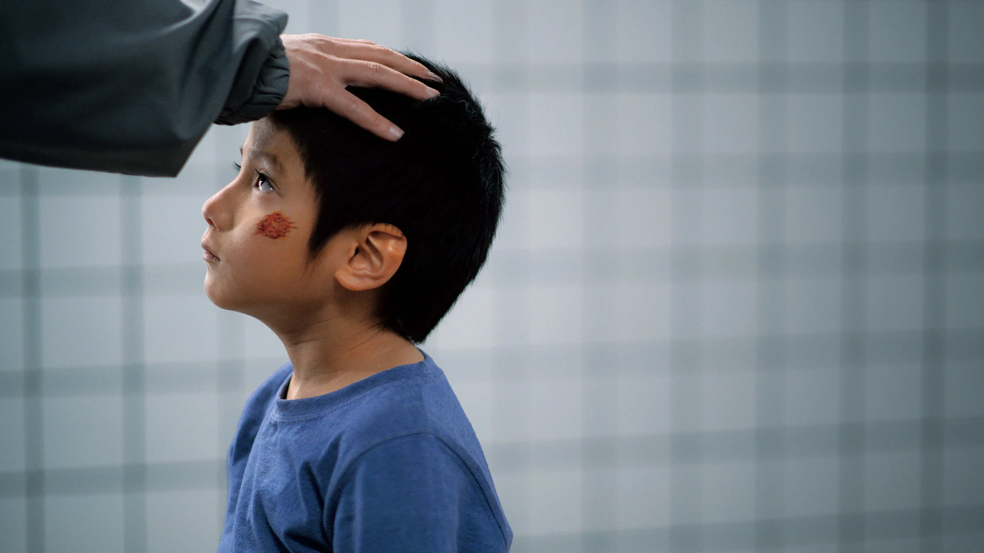 A boy in a blue shirt looks up as an adult rests a hand on his head.