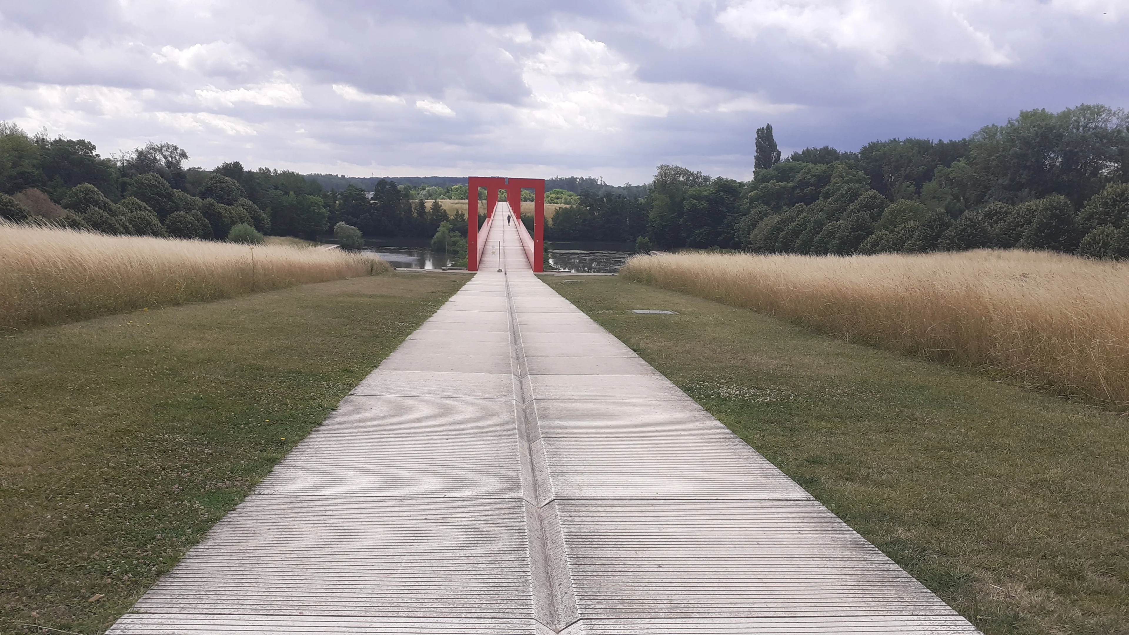 A long white walkway leads through a red gateway toward the tree-lined Oise valley in Cergy.