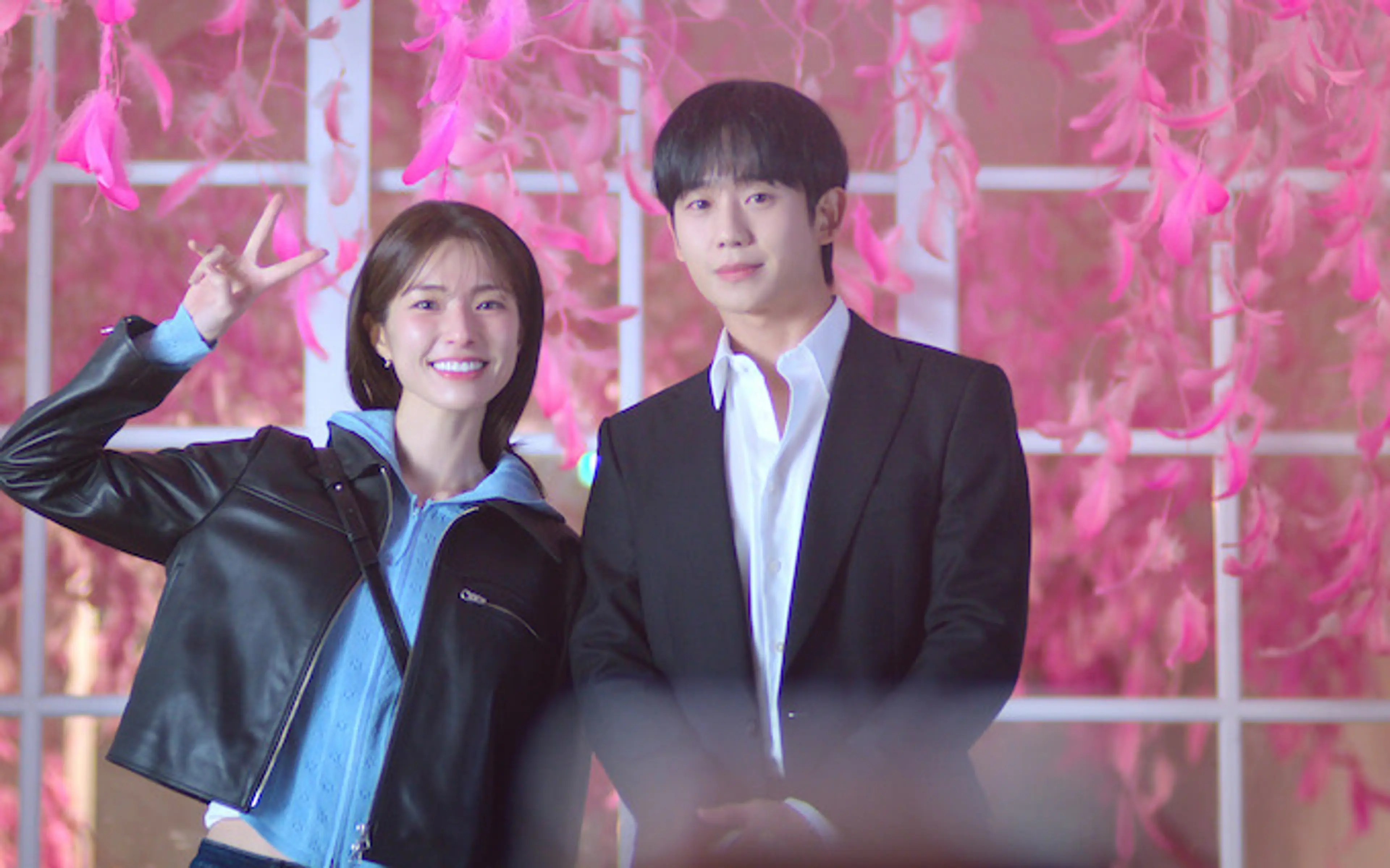 Ha Young smiles and makes a peace sign beside Jung Hae-in in a dark suit, with pink flowers behind them.