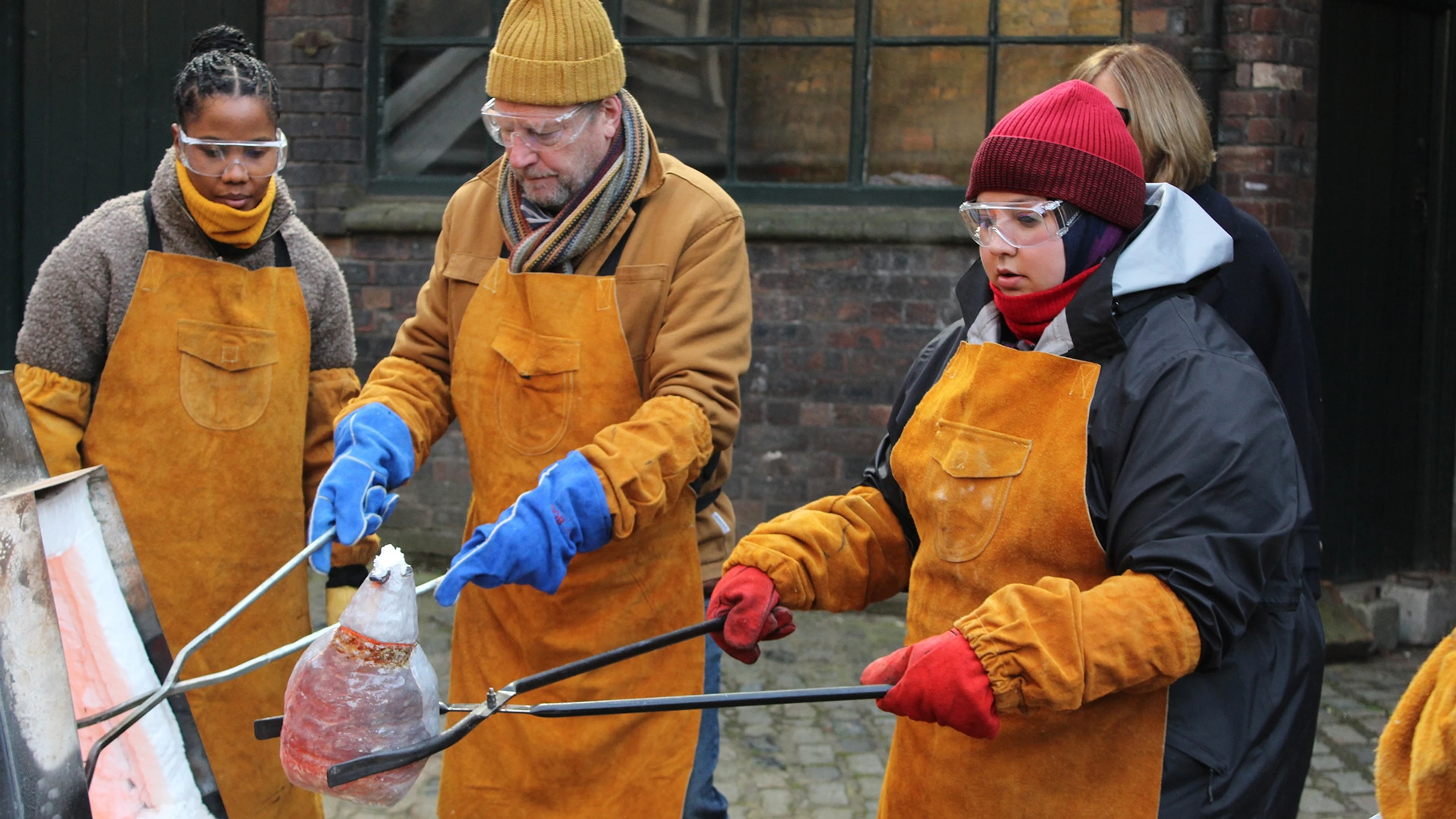 Celebrity potters in protective aprons and goggles work with hot Raku ceramics outdoors.
