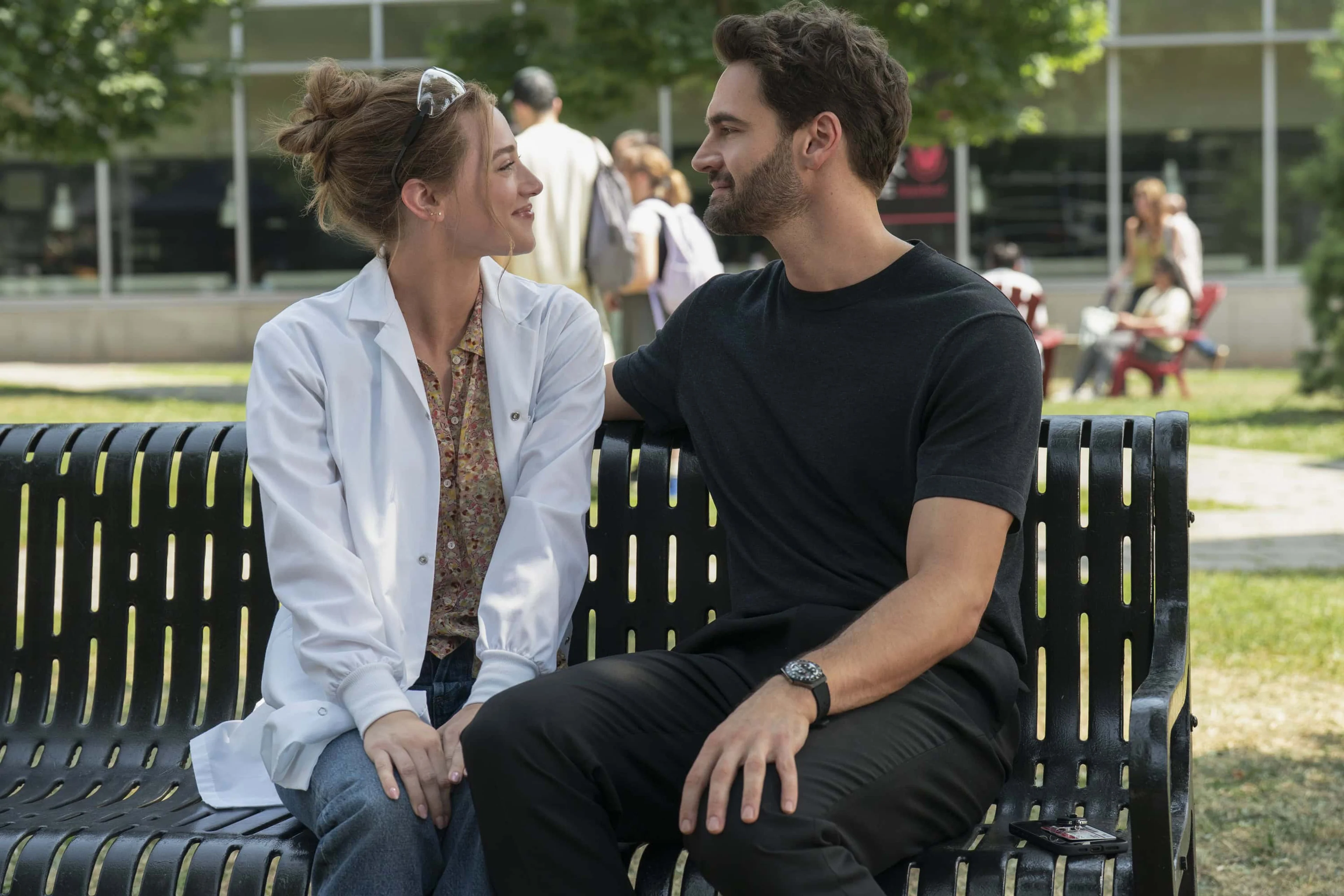 Olive and Adam smile at each other while sitting on a campus bench.