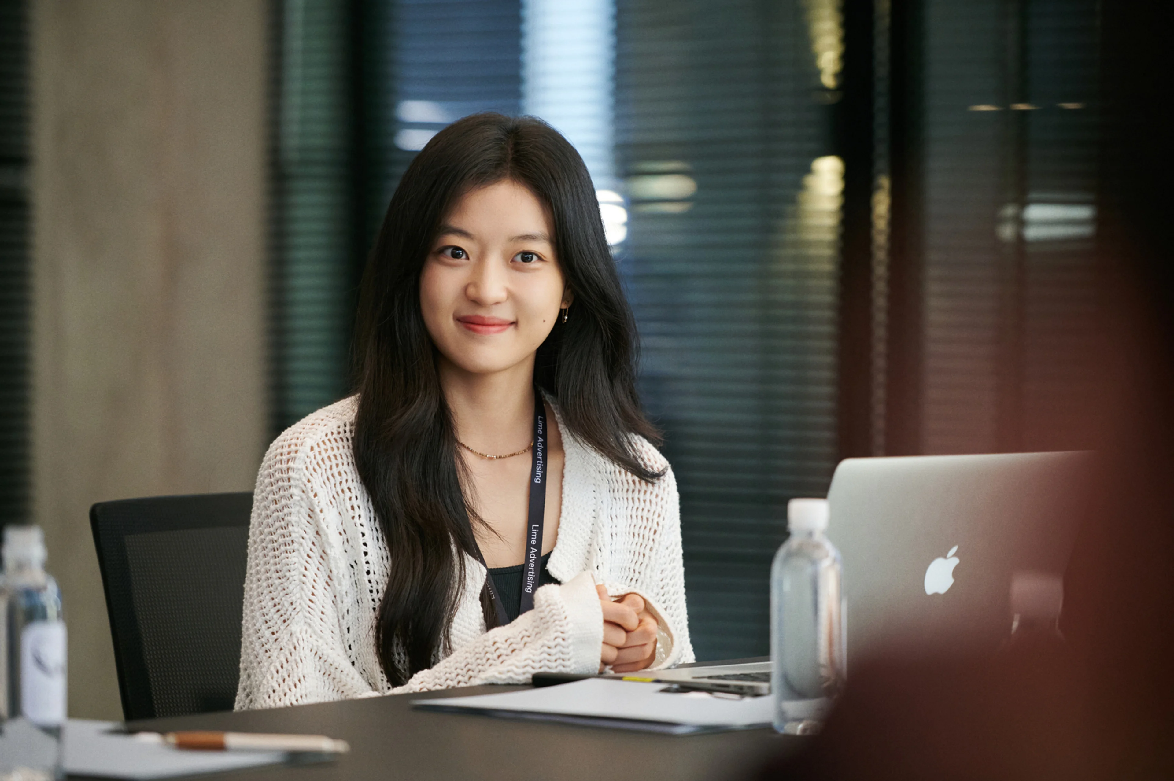 Sun Qian smiles at an open laptop on her desk while wearing a white cardigan and lanyard.