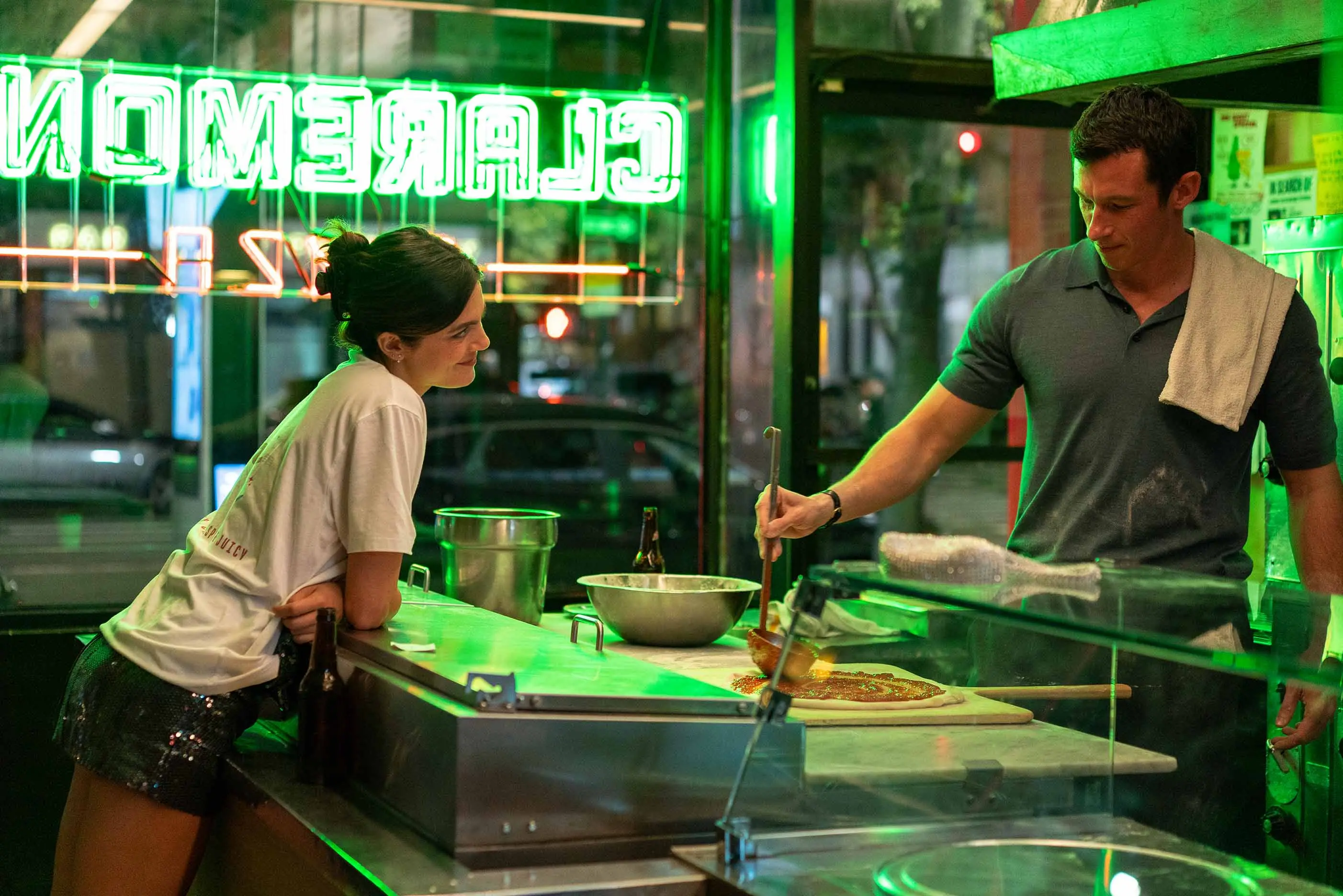 Monica Barbaro as Allie watches Callum Turner as Owen make pizza behind a neon-lit counter in One Night Only.