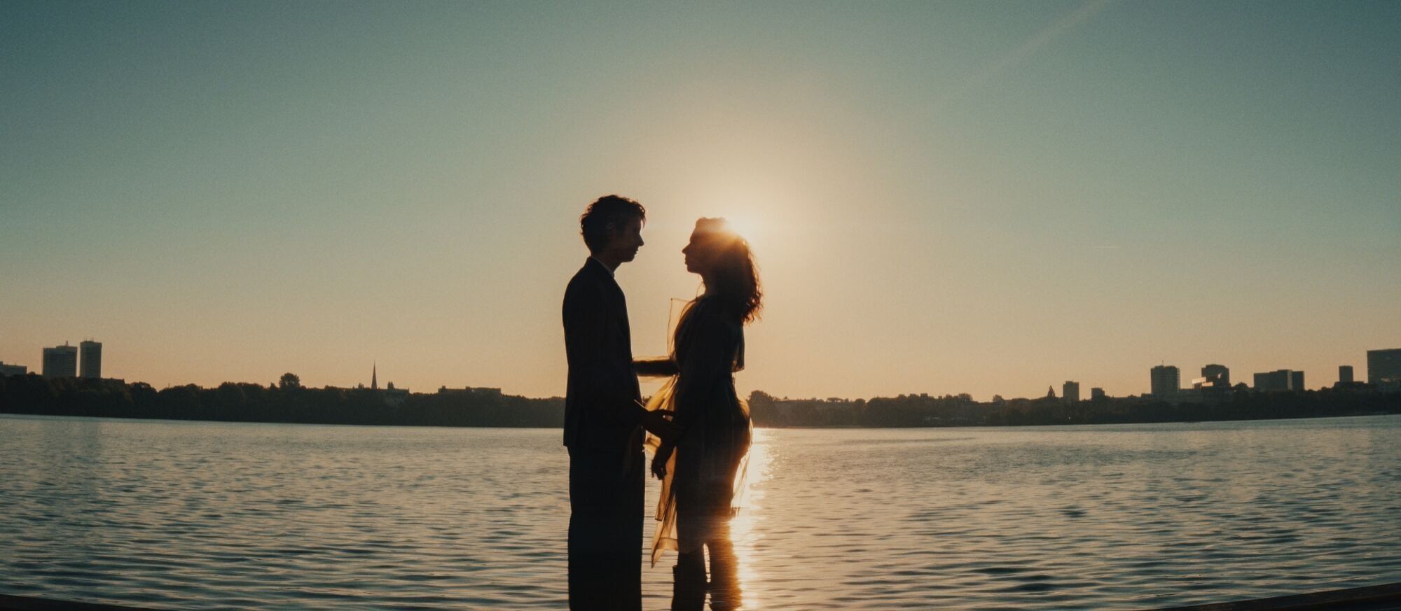 A couple stands together in shallow water at sunset with a city skyline behind them.