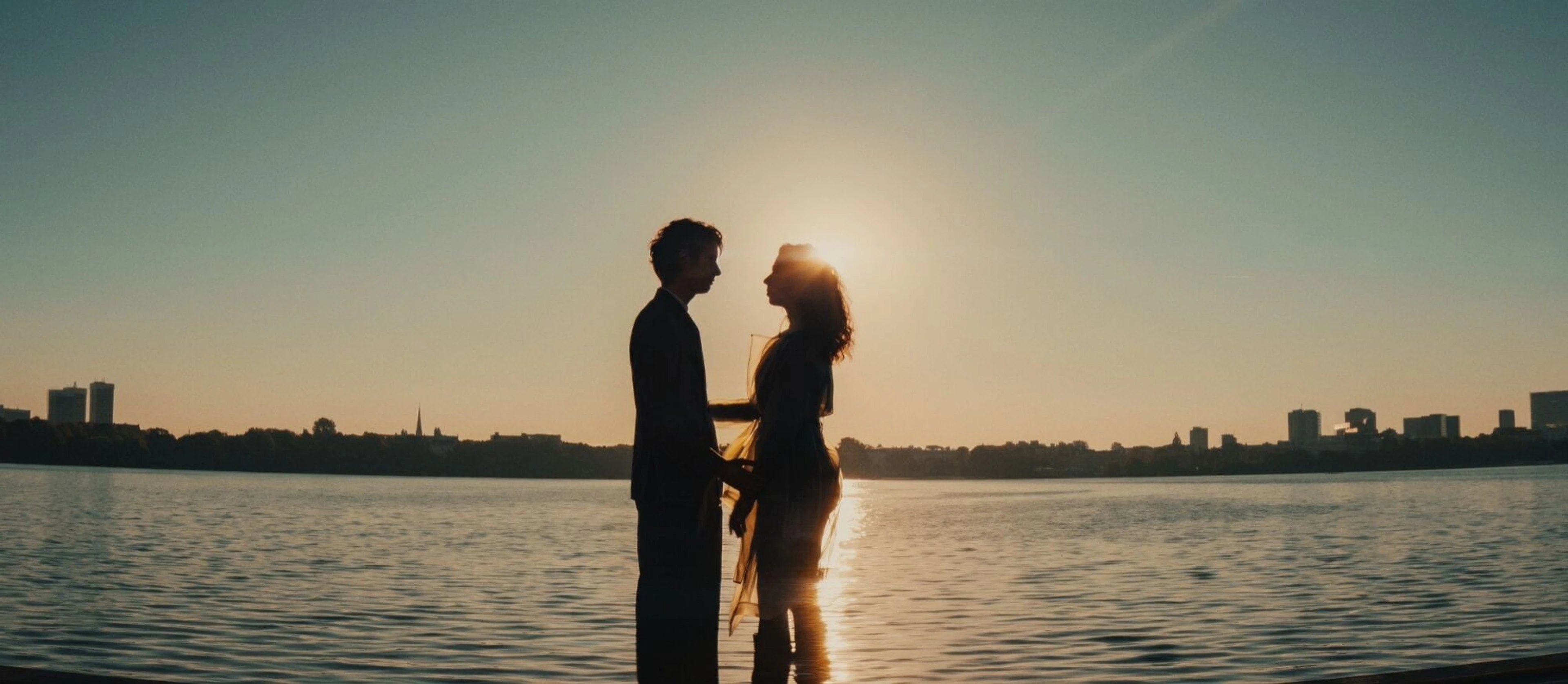 A couple stands together in shallow water at sunset with a city skyline behind them.