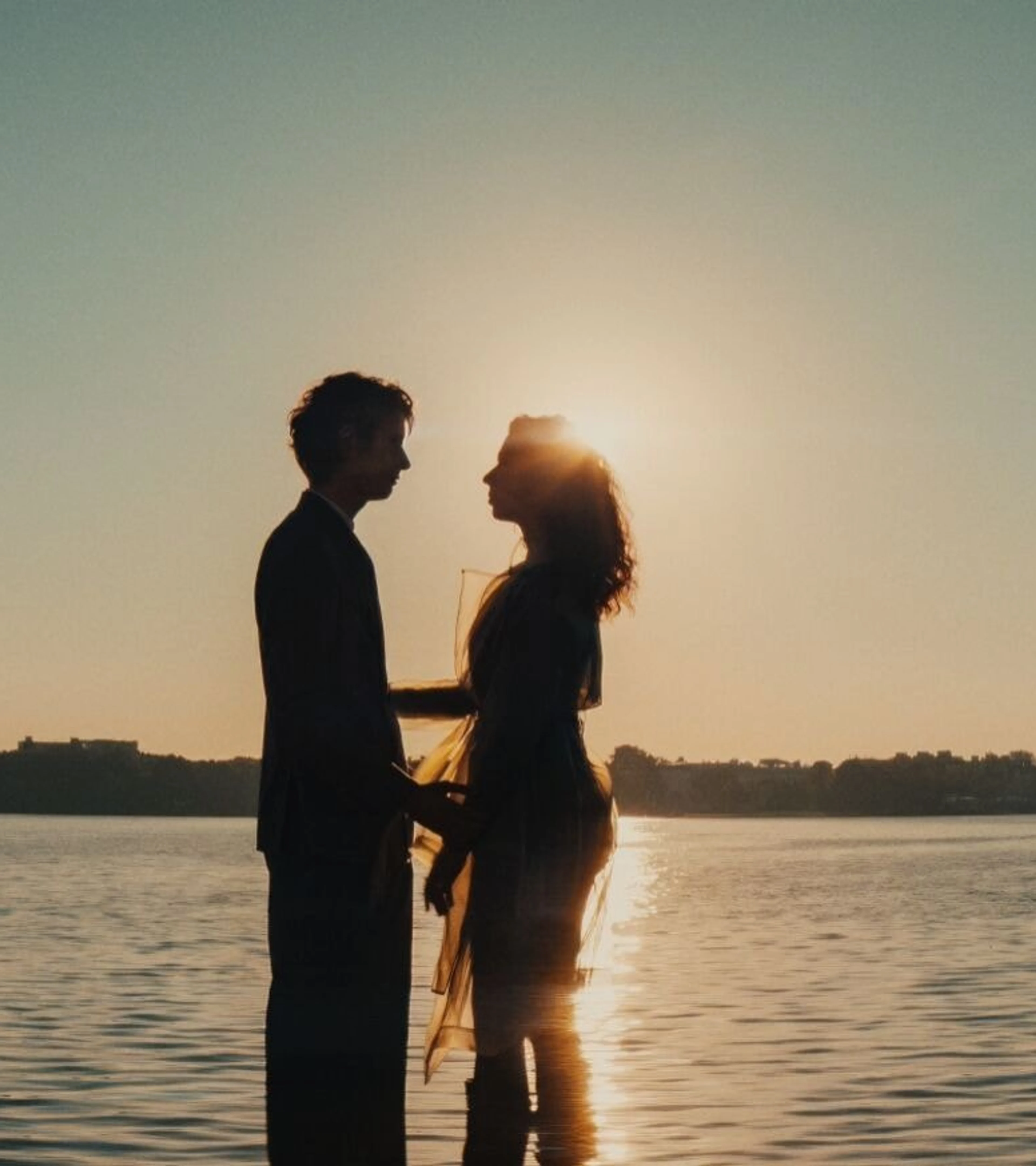 A couple stands together in shallow water at sunset with a city skyline behind them.