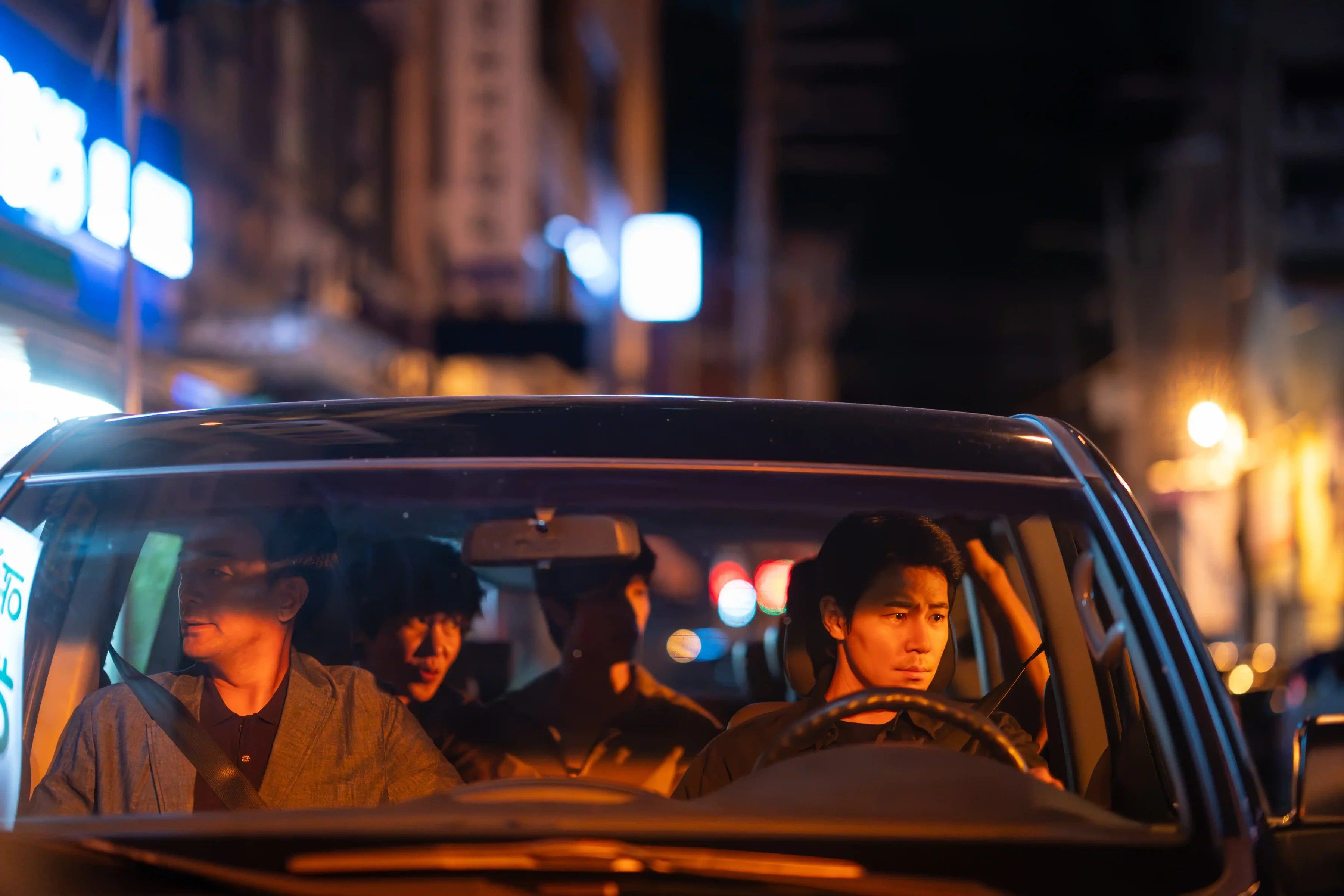 Three men ride in a car at night, seen through the windshield.