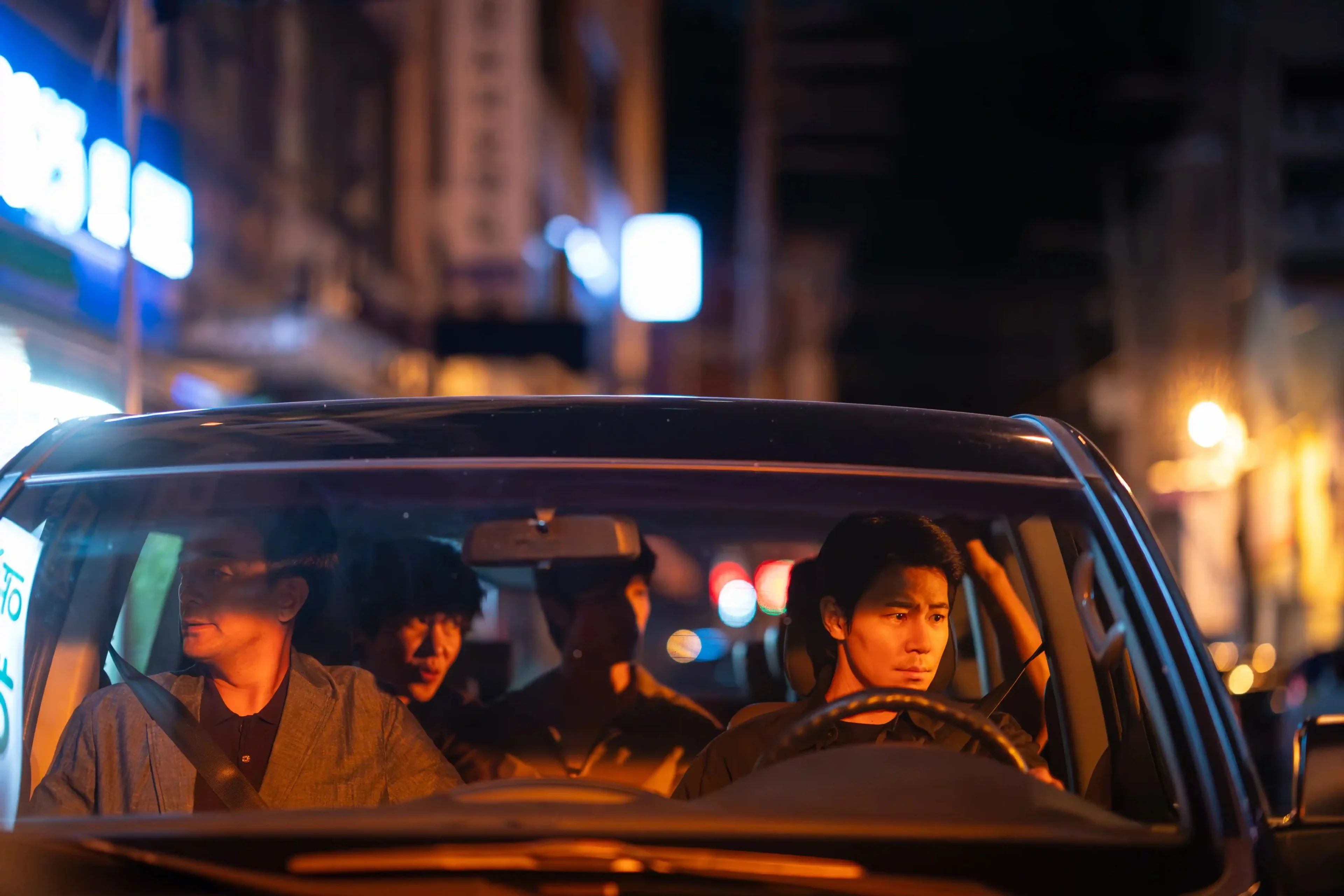 Three men ride in a car at night, seen through the windshield.