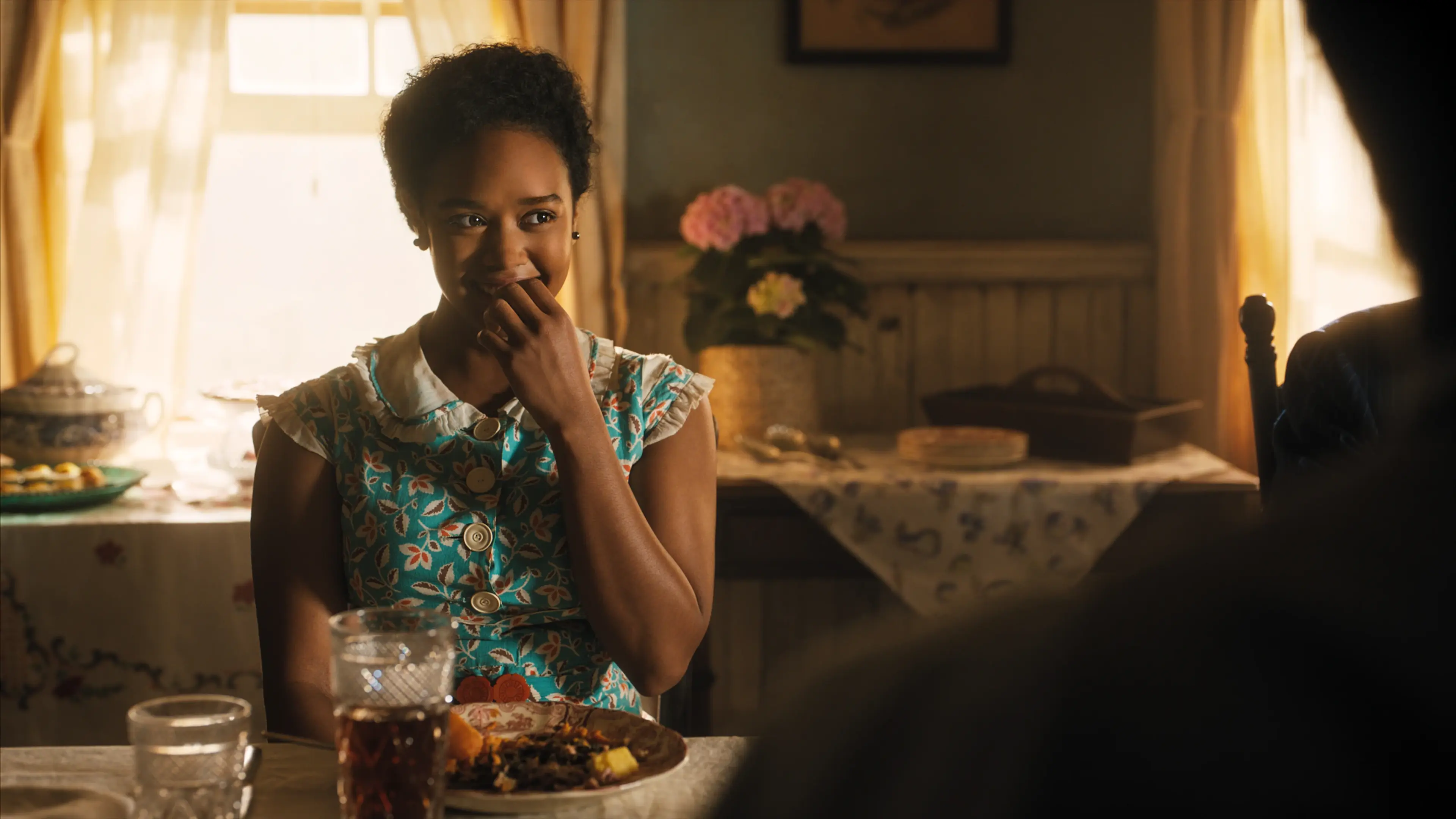 A woman in a floral dress sits at a dining table in The Color Purple (2023).