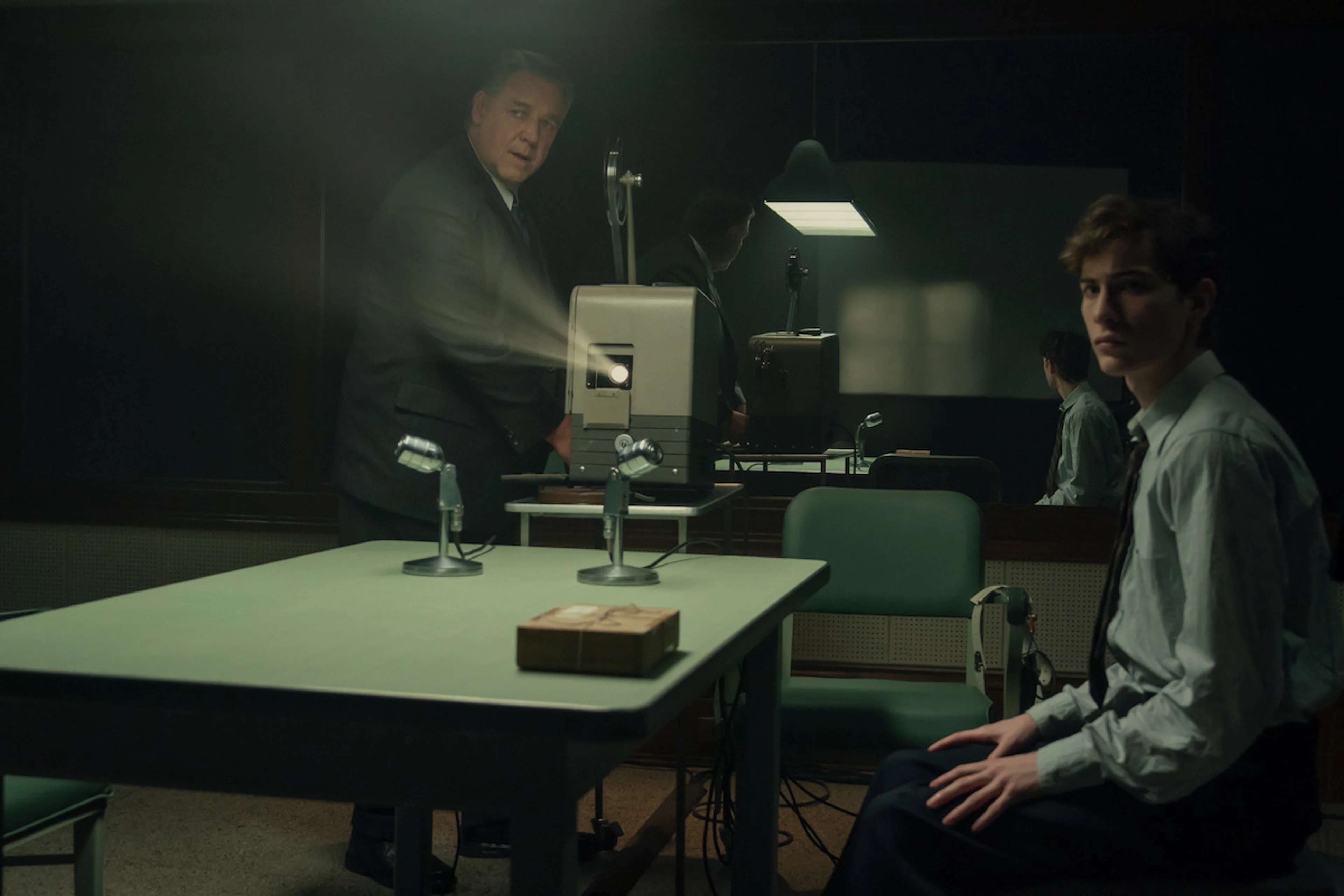 Henry Murray stands beside a projector while Ted Kaczynski sits at a table fitted with microphones.