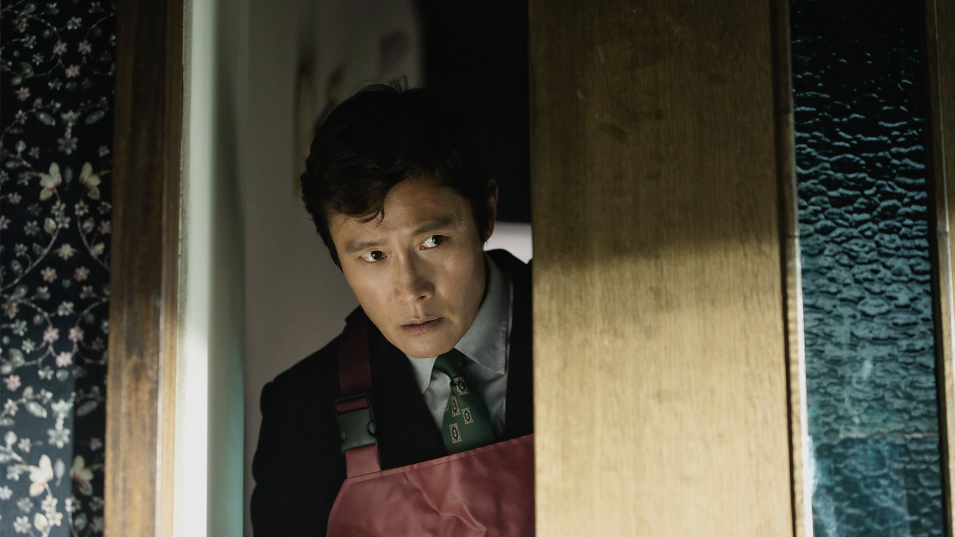 Lee Byung-hun as Man-su peers through a partly open wooden door, wearing a red apron.