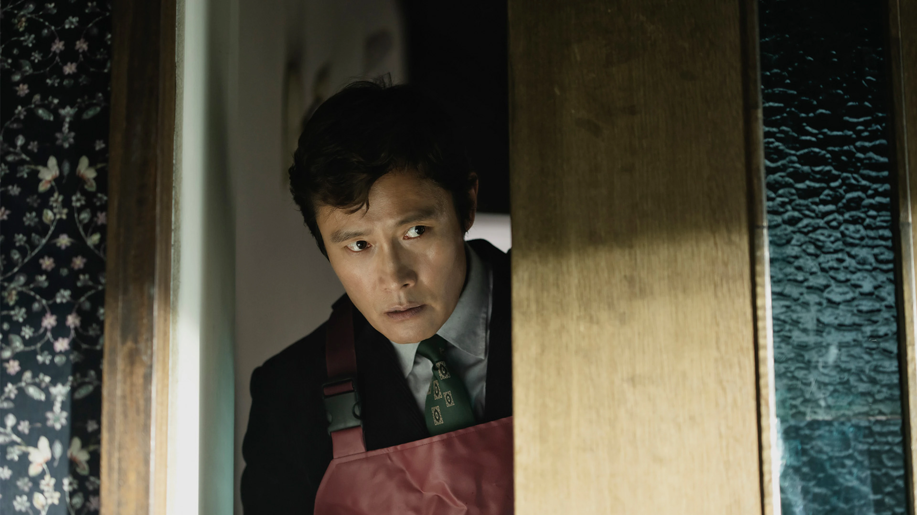 Lee Byung-hun as Man-su peers through a partly open wooden door, wearing a red apron.