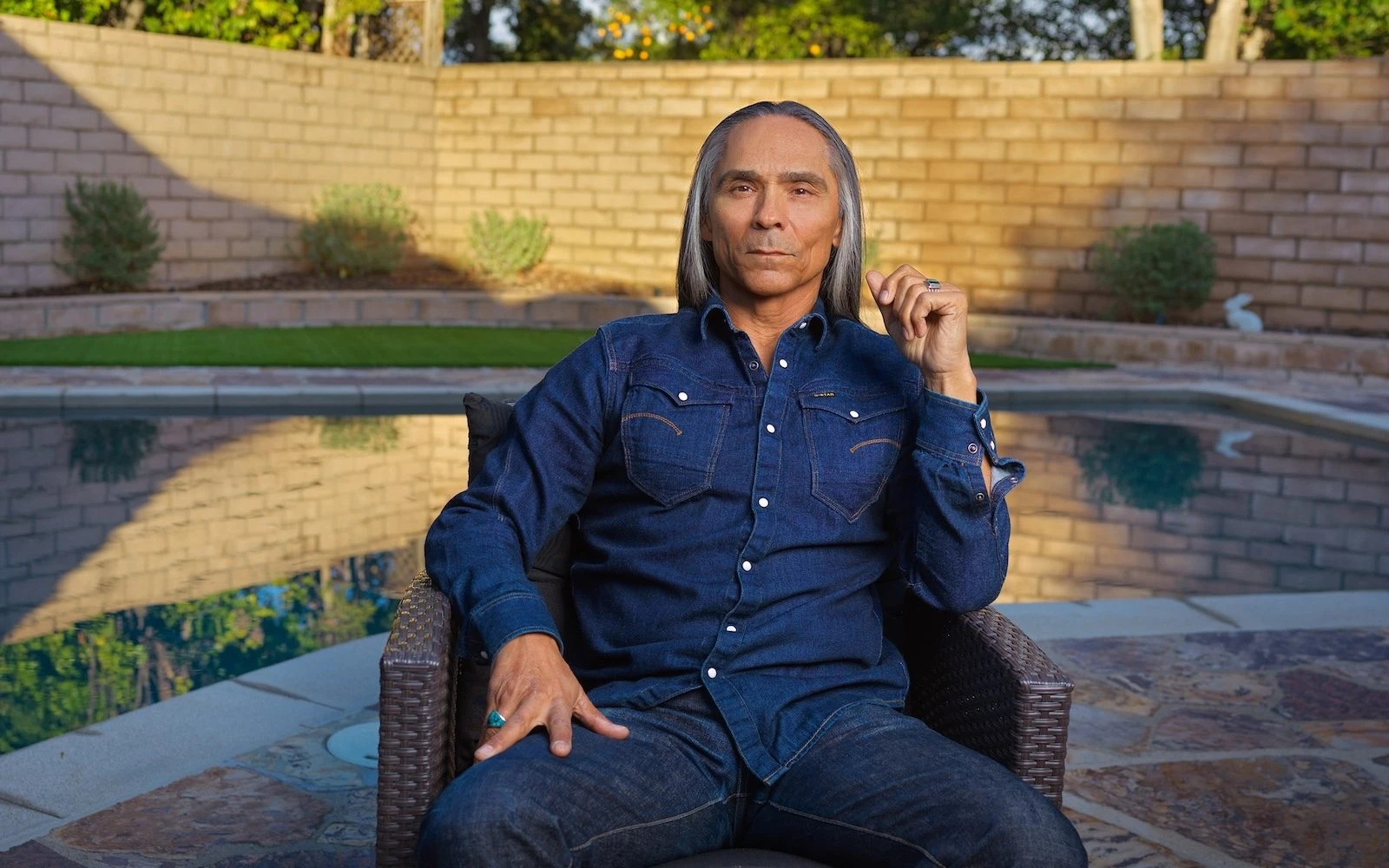 Zahn McClarnon wearing a denim shirt and seated beside a swimming pool.