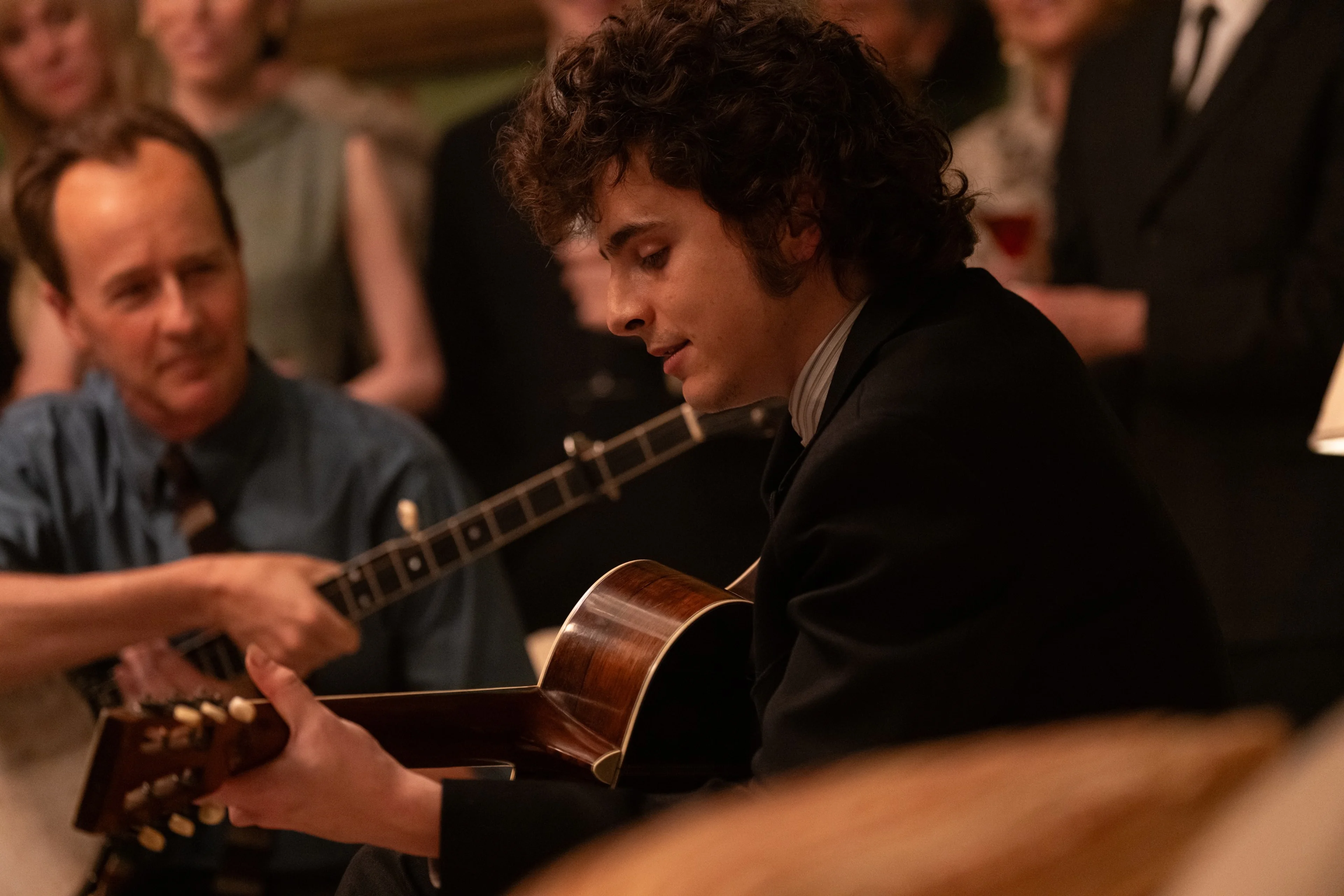 Edward Norton with a banjo and Timothée Chalamet with a guitar sit among listeners.