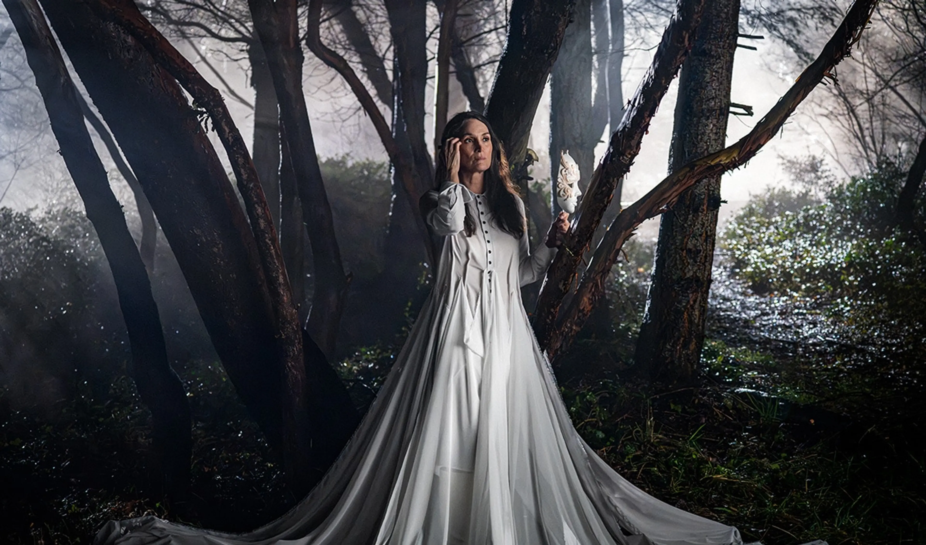 Carrie-Anne Moss as Katherine stands in a forest in a white gown, holding a dark mask.