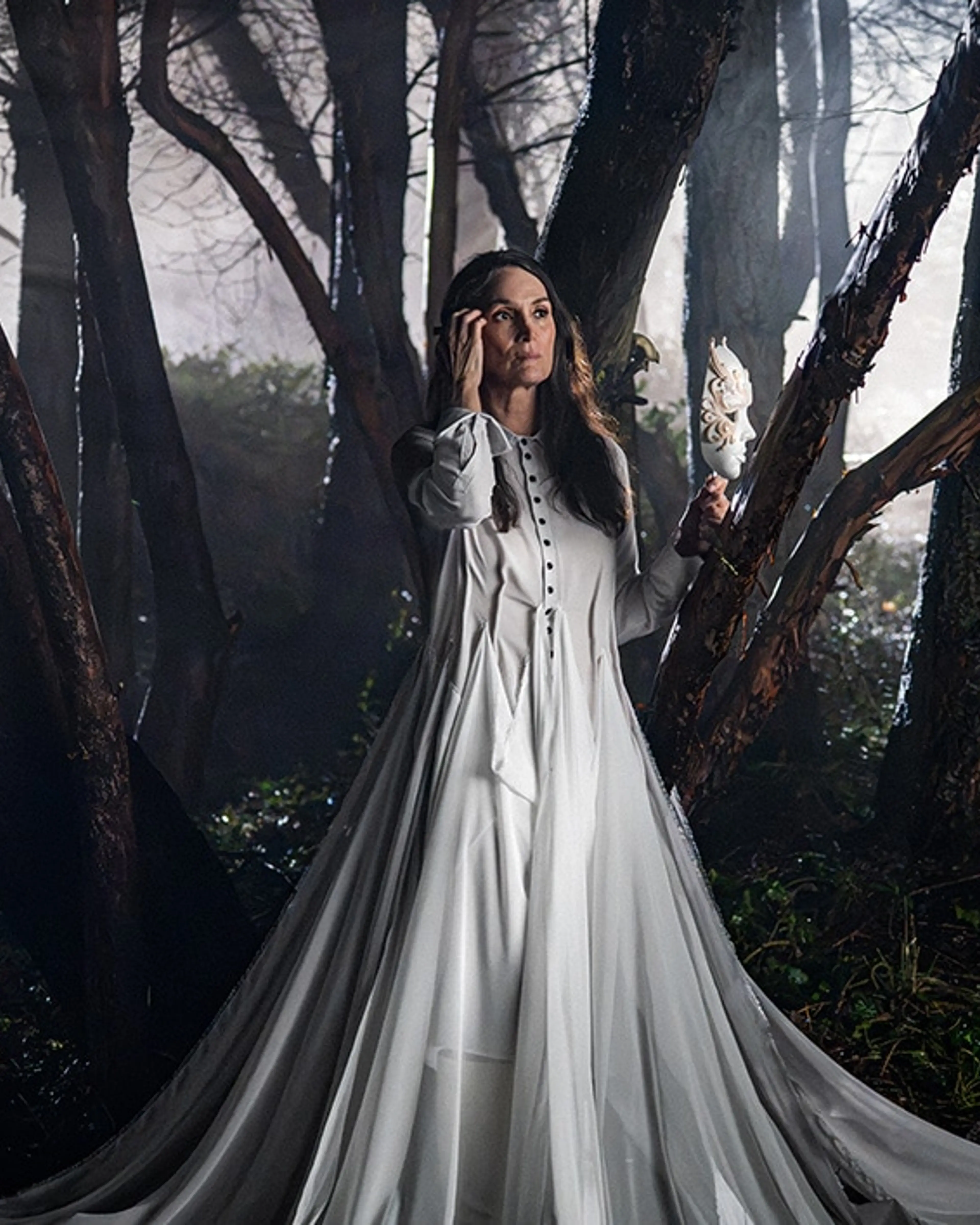 Carrie-Anne Moss as Katherine stands in a forest in a white gown, holding a dark mask.