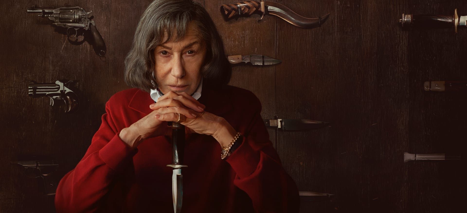 Helen Mirren as Patricia Highsmith sits in red before a wall of knives and guns.