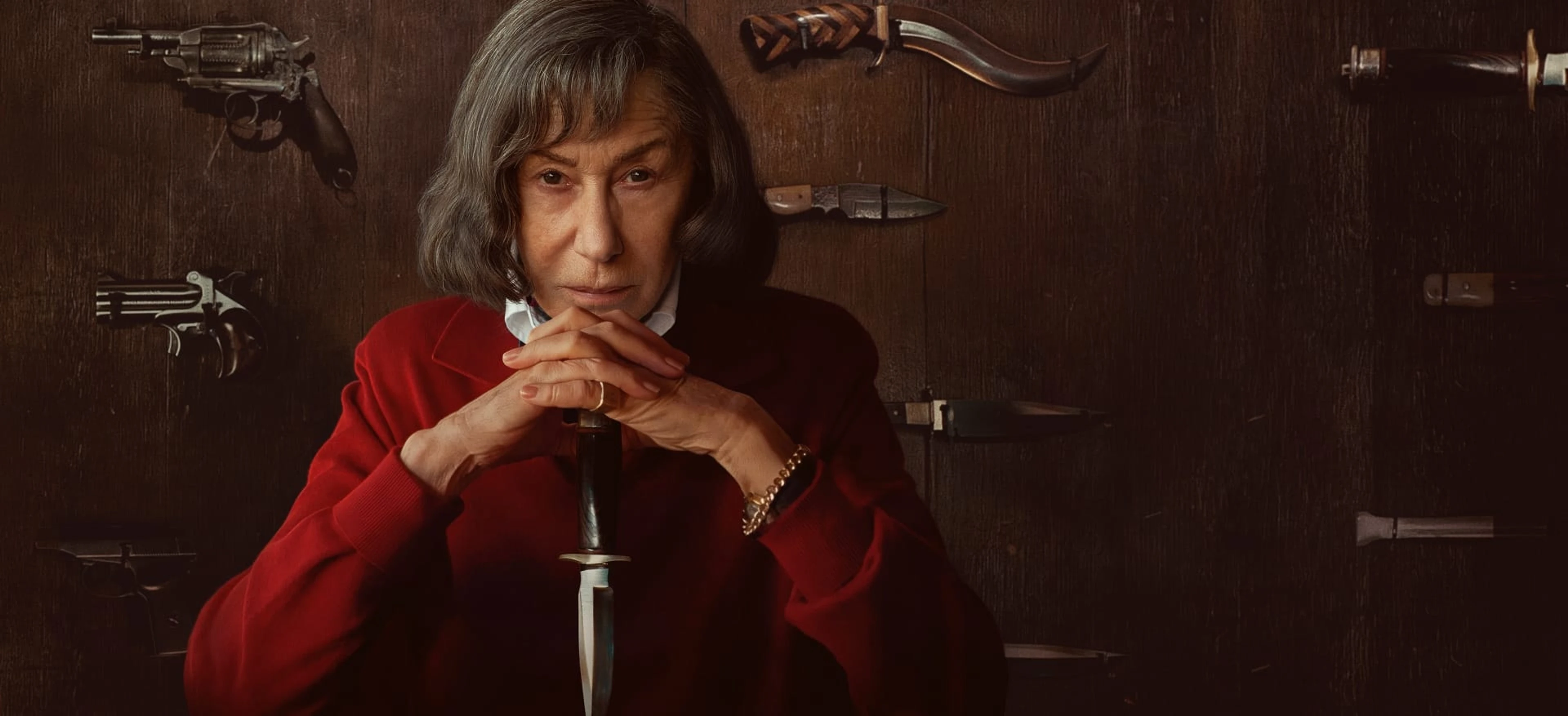 Helen Mirren as Patricia Highsmith sits in red before a wall of knives and guns.