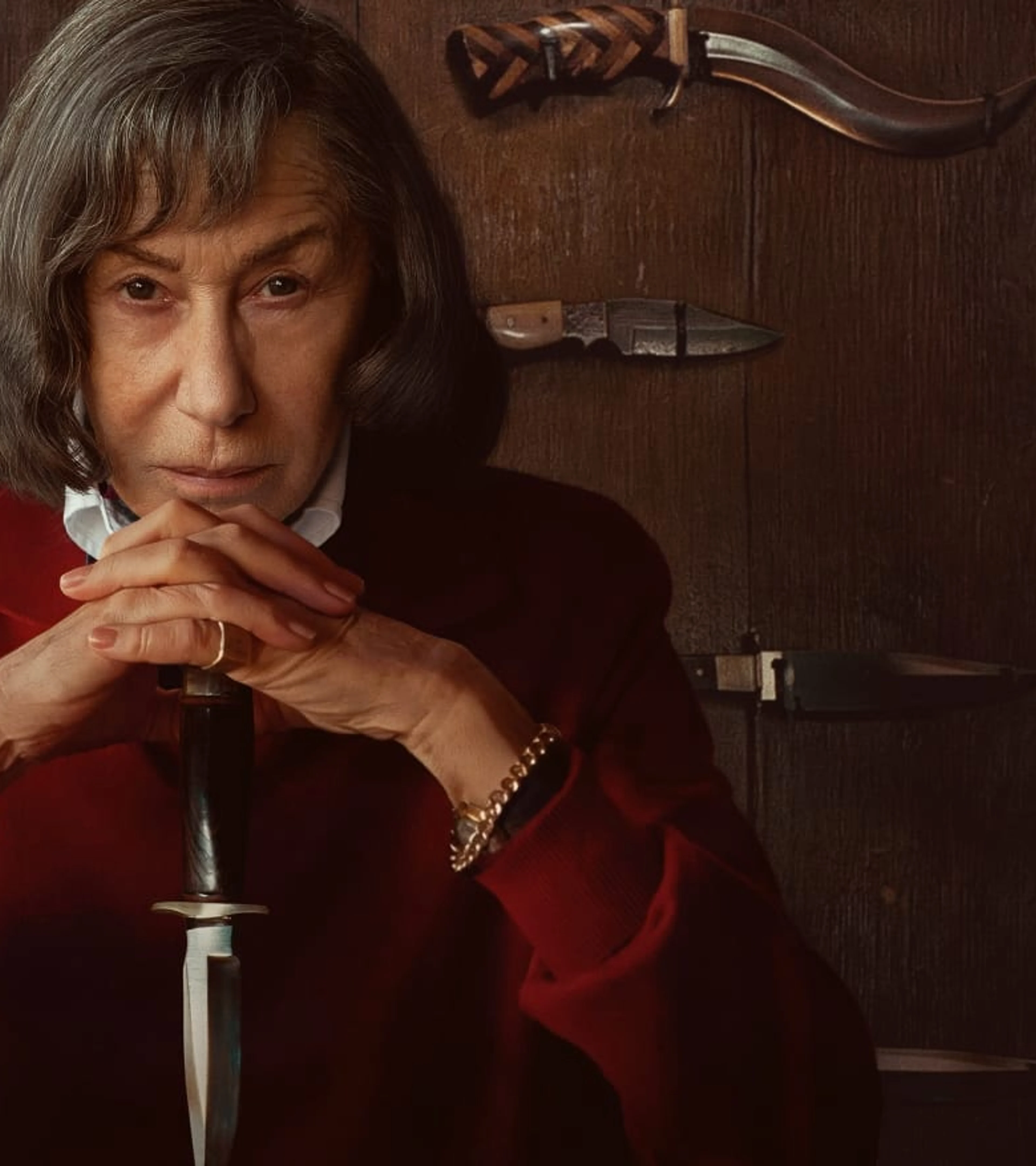 Helen Mirren as Patricia Highsmith sits in red before a wall of knives and guns.