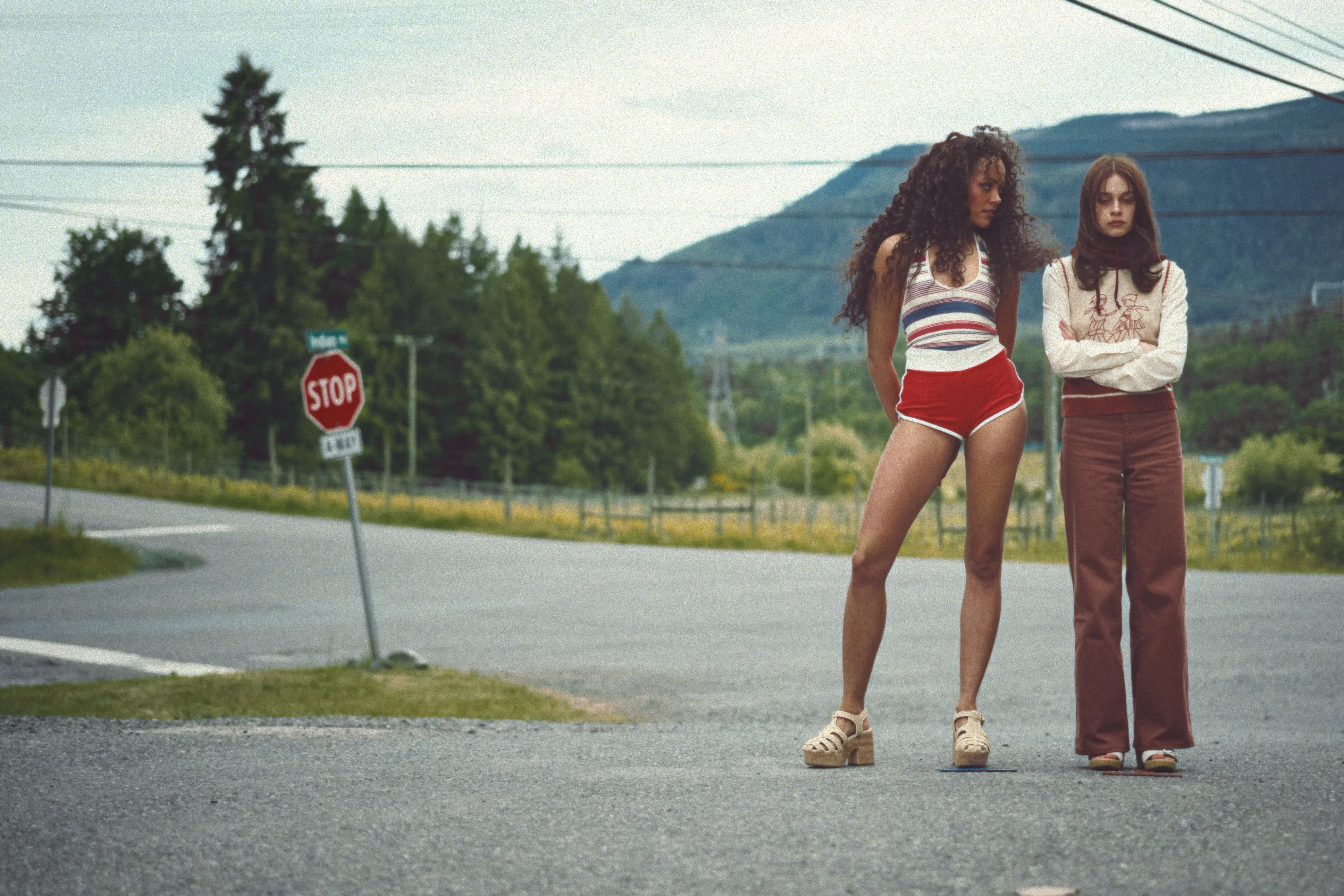 Two young women stand beside a vehicle on a road through the woods.