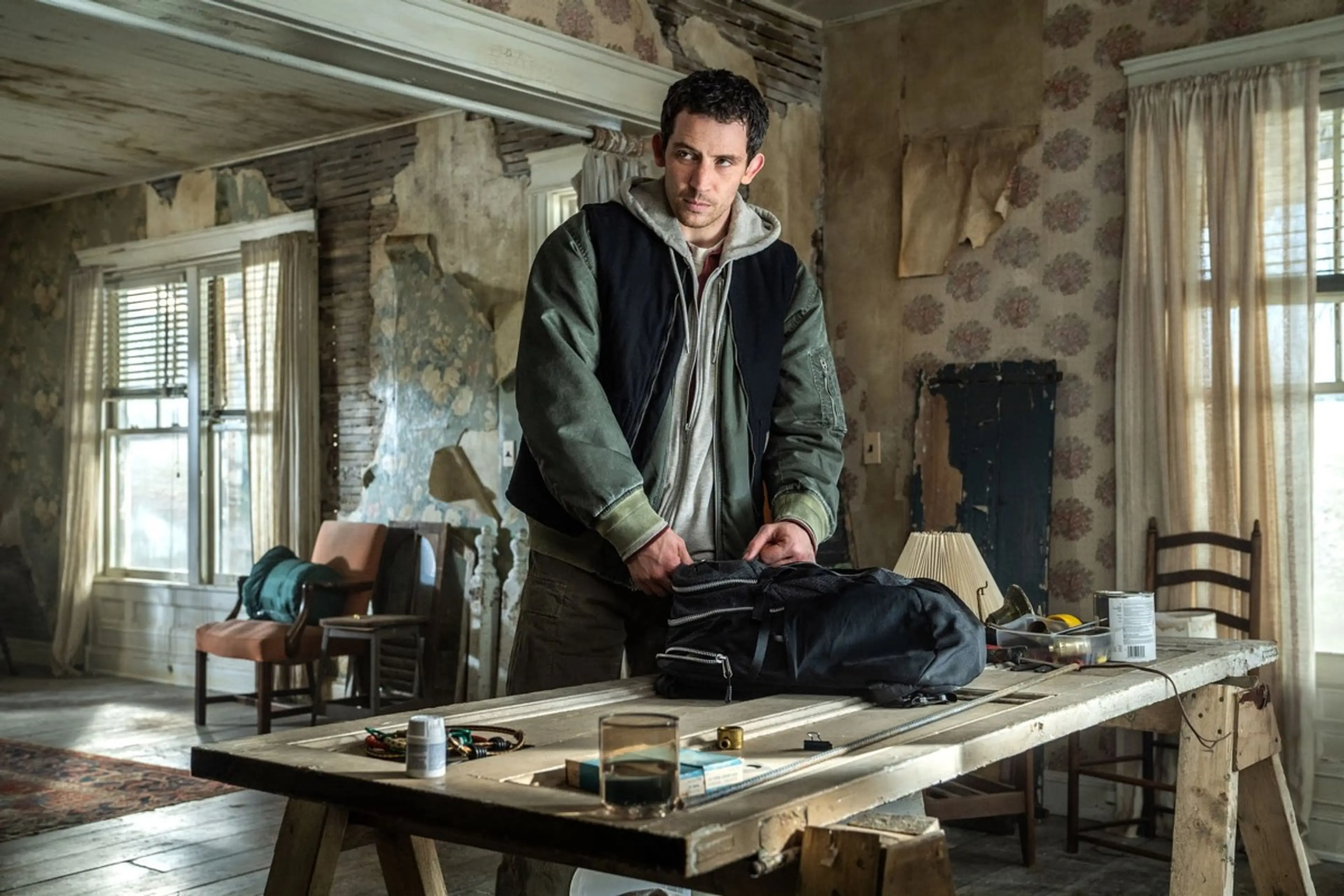 Josh O’Connor as Daniel stands behind a bag on a table in a worn room.