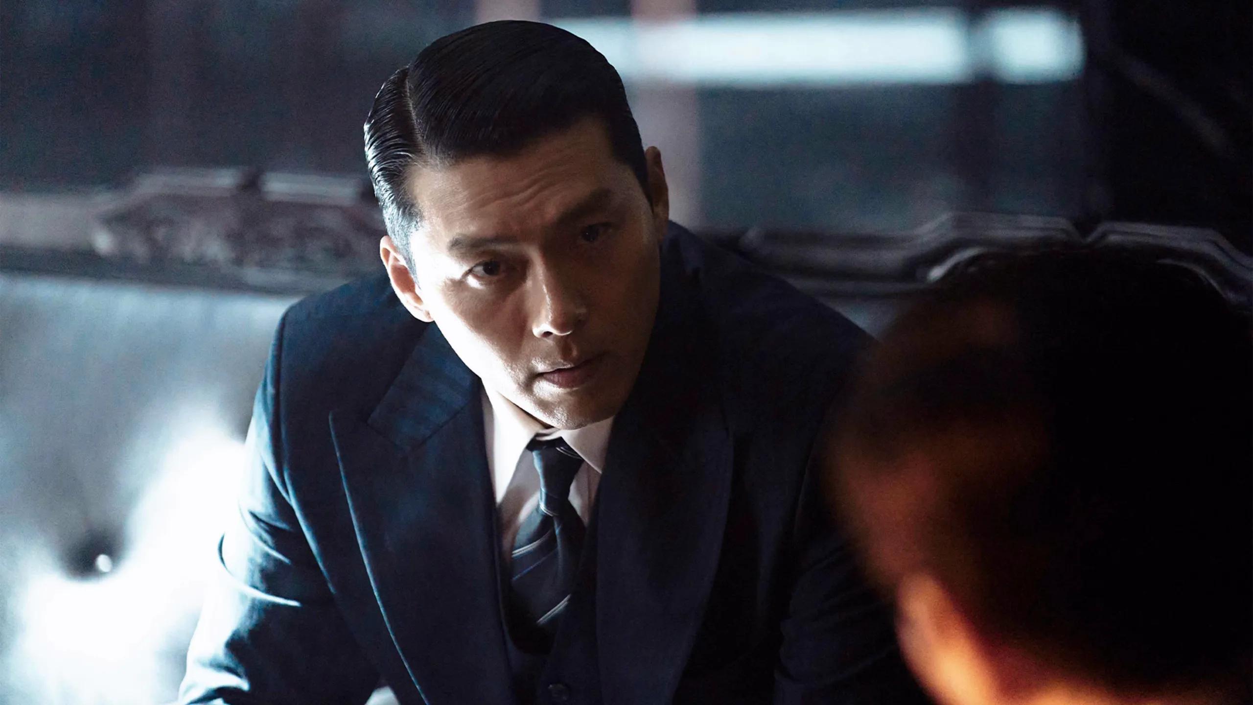 Hyun Bin as Ki-tae leans forward in a dark blue suit and looks toward someone opposite him.