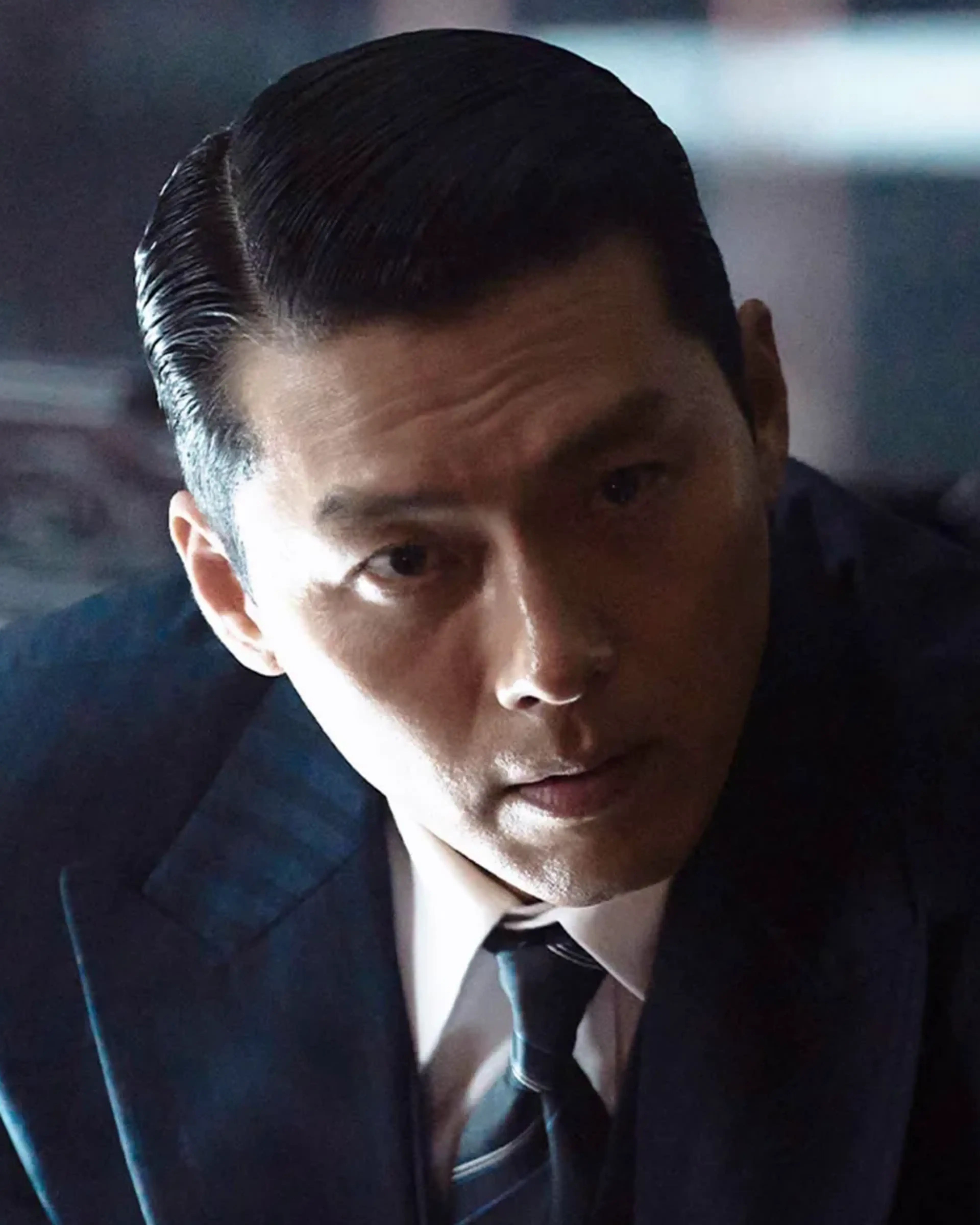 Hyun Bin as Ki-tae leans forward in a dark blue suit and looks toward someone opposite him.