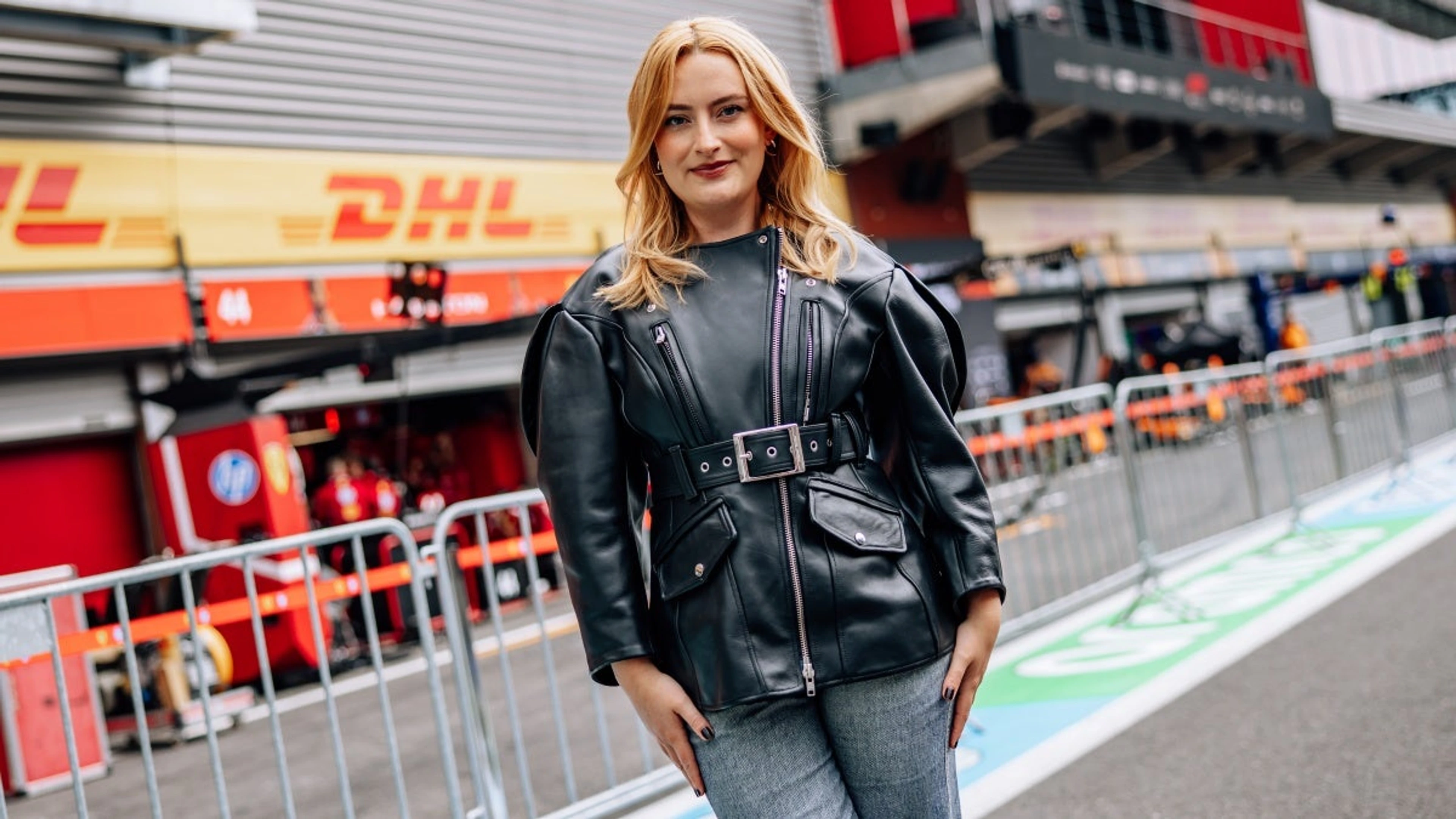 Amelia Dimoldenberg stands in front of an F1 paddock wearing a black leather jacket.