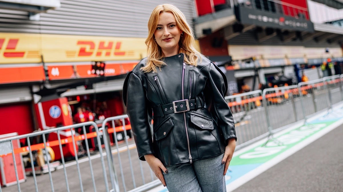 Amelia Dimoldenberg stands in front of an F1 paddock wearing a black leather jacket.