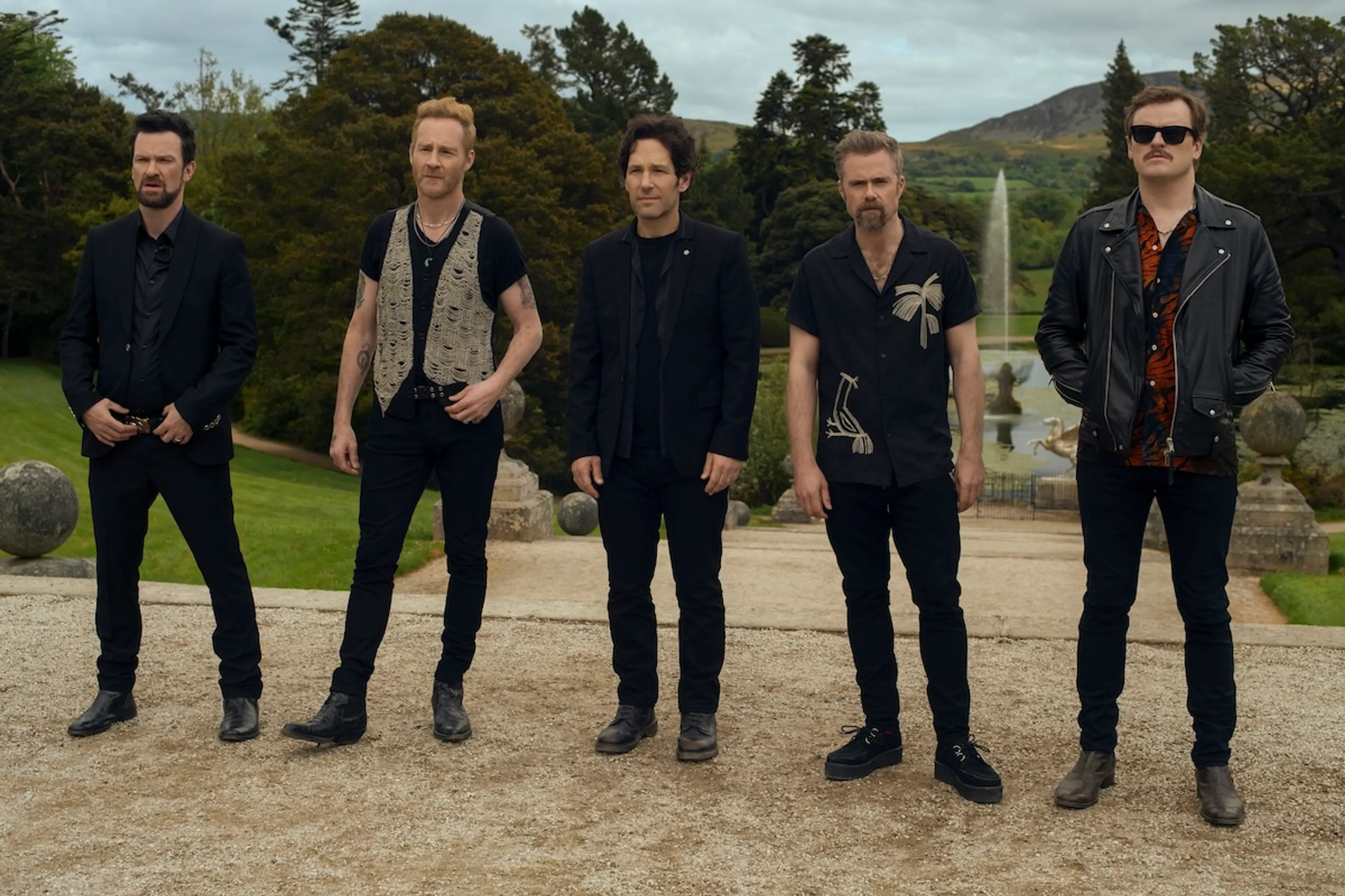 Rick stands with four bandmates on a gravel path in a garden.