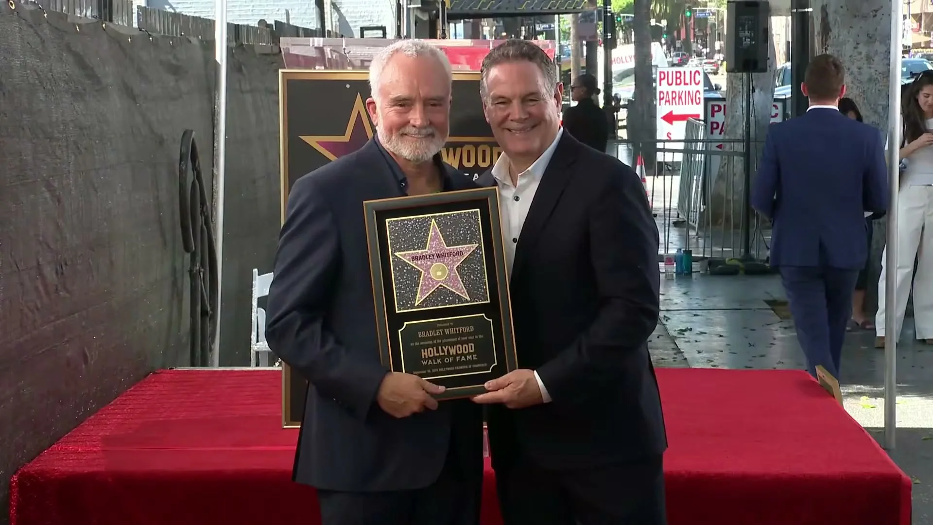 Bradley Whitford and a man in a dark suit hold a framed Hollywood Walk of Fame plaque and face the camera.