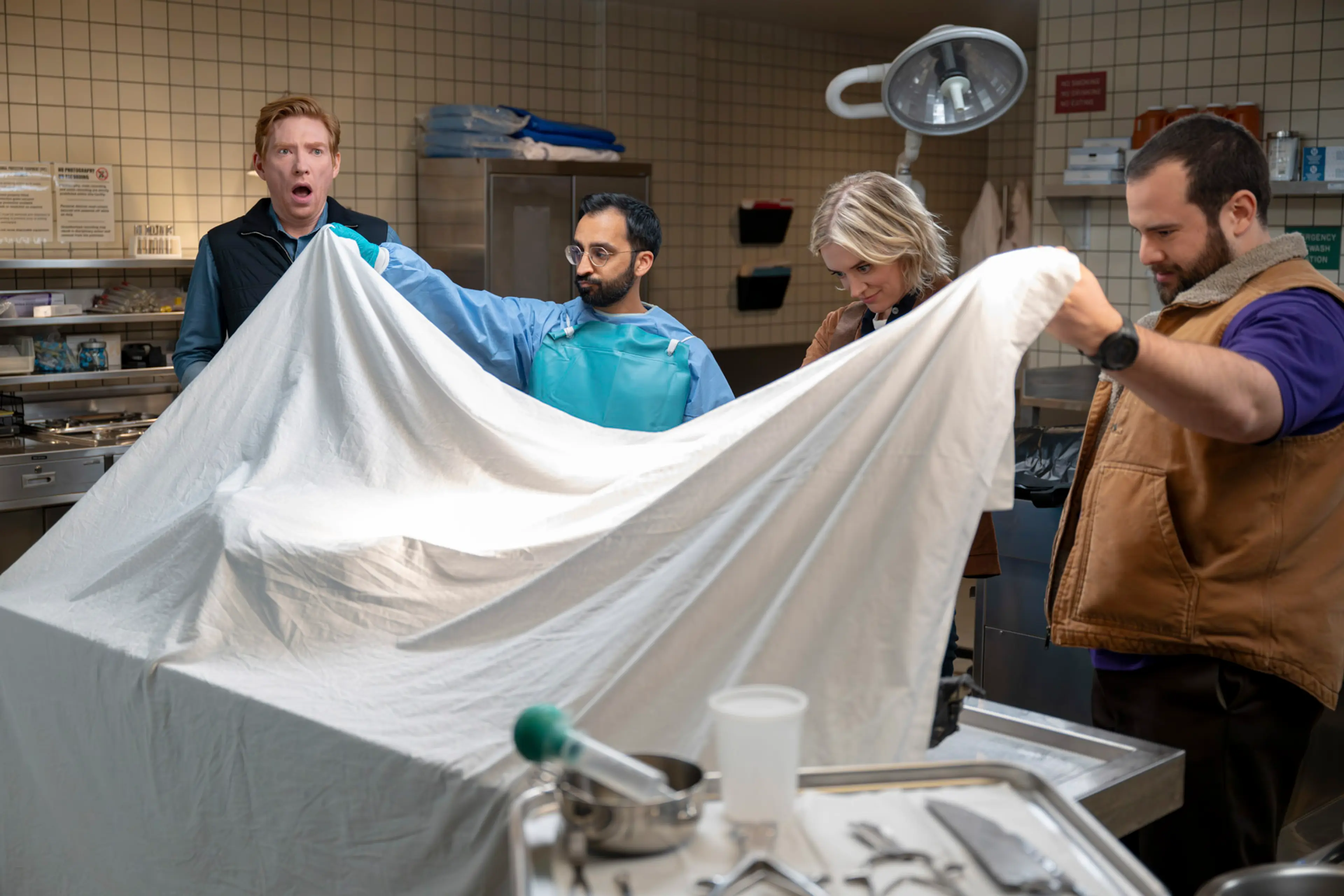 Ned, Mare and Travis help lift a sheet from a covered body in a medical room.