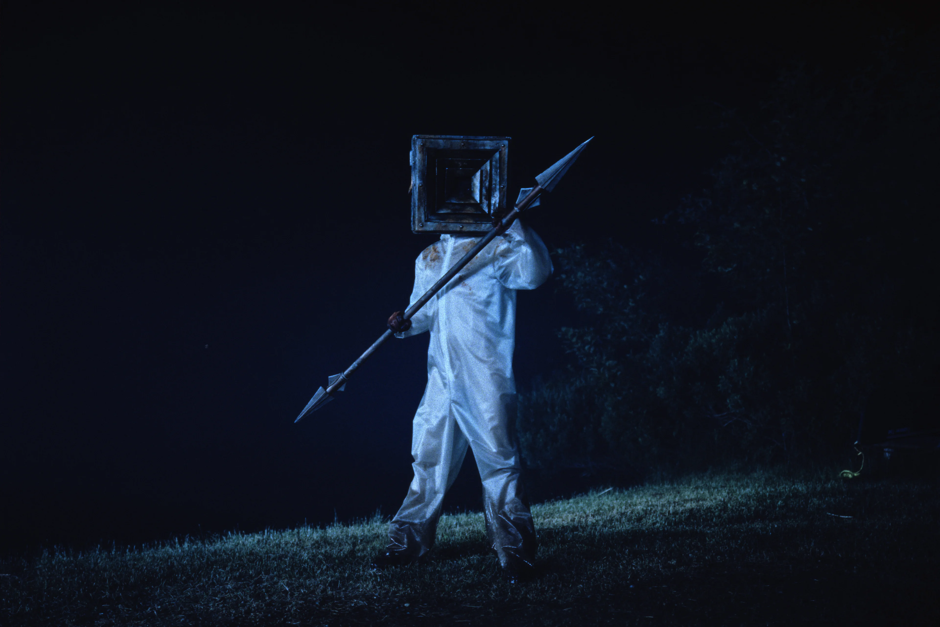 Little Death stands outdoors at night wearing a square mask and a clear covering over white clothes, holding a long weapon.