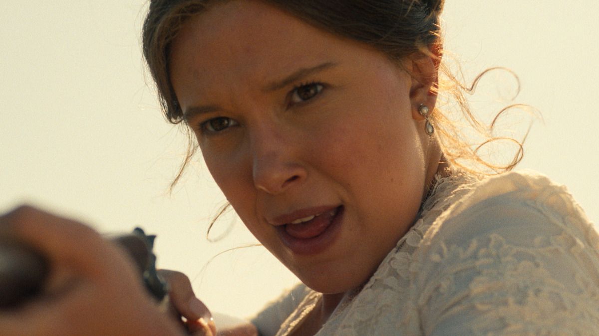 Millie Bobby Brown as Enola Holmes aims a gun outdoors, wearing a white lace dress.