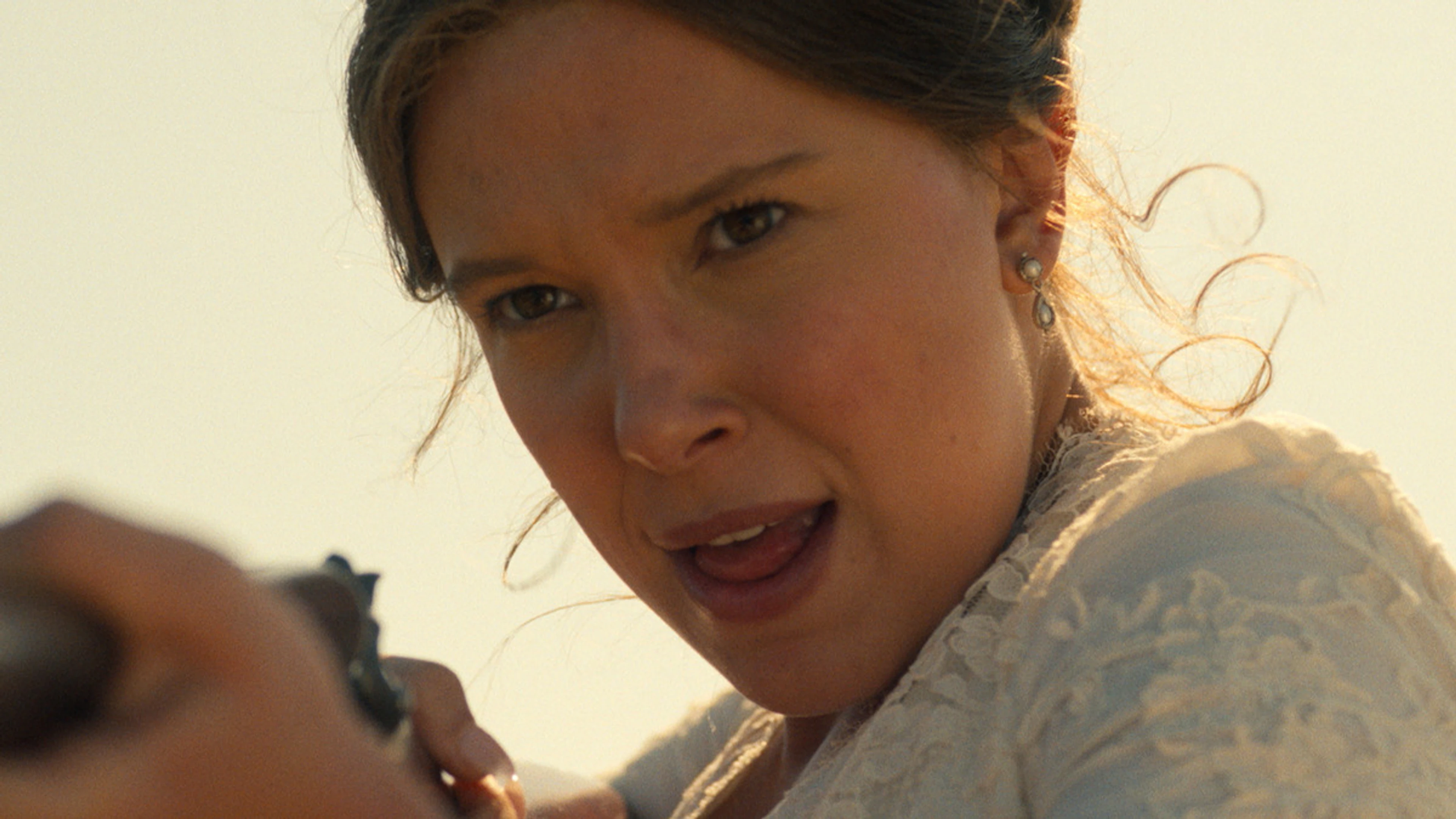 Millie Bobby Brown as Enola Holmes aims a gun outdoors, wearing a white lace dress.