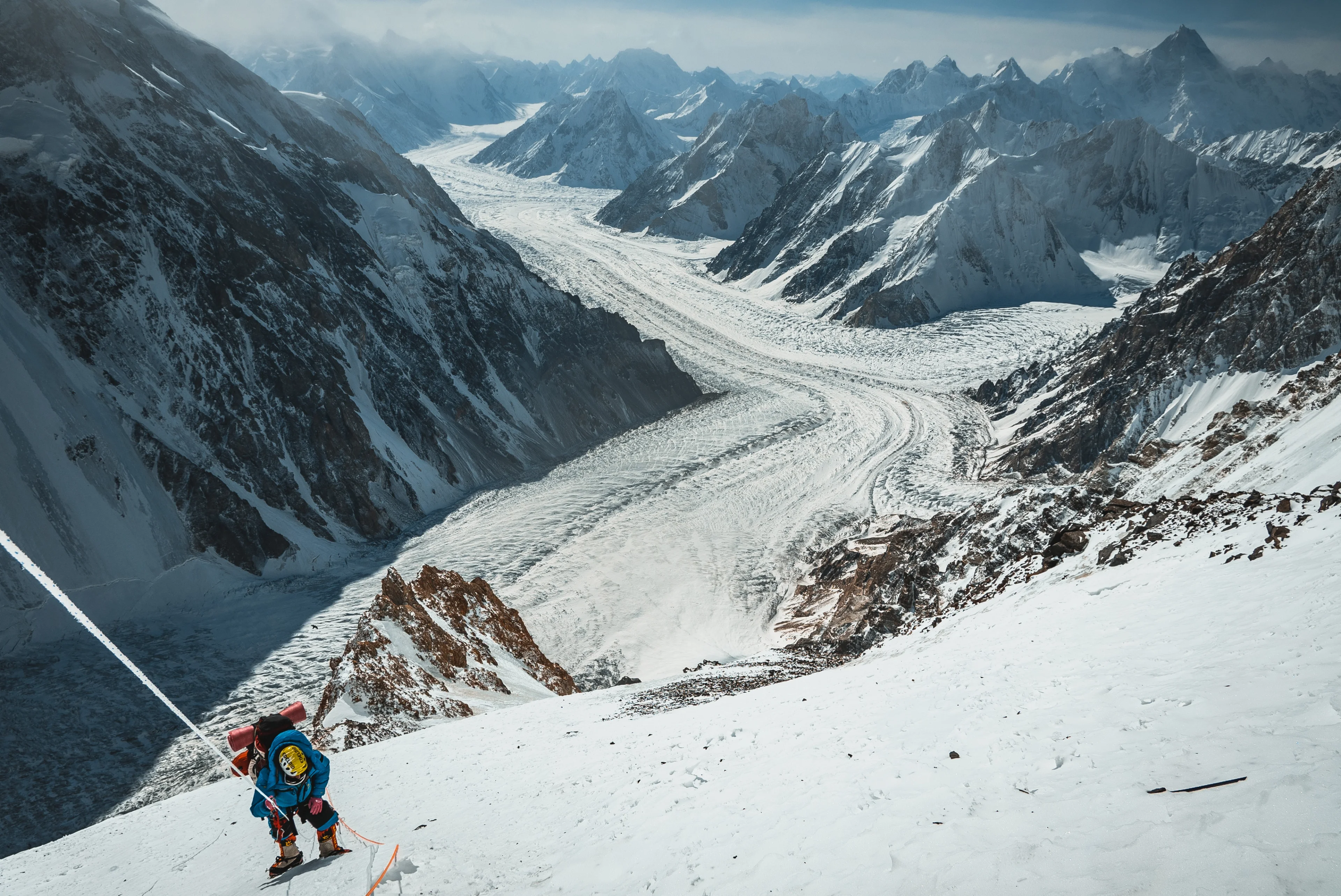 A climber in a blue suit ascends a steep snowy slope with a glacier and mountains below.