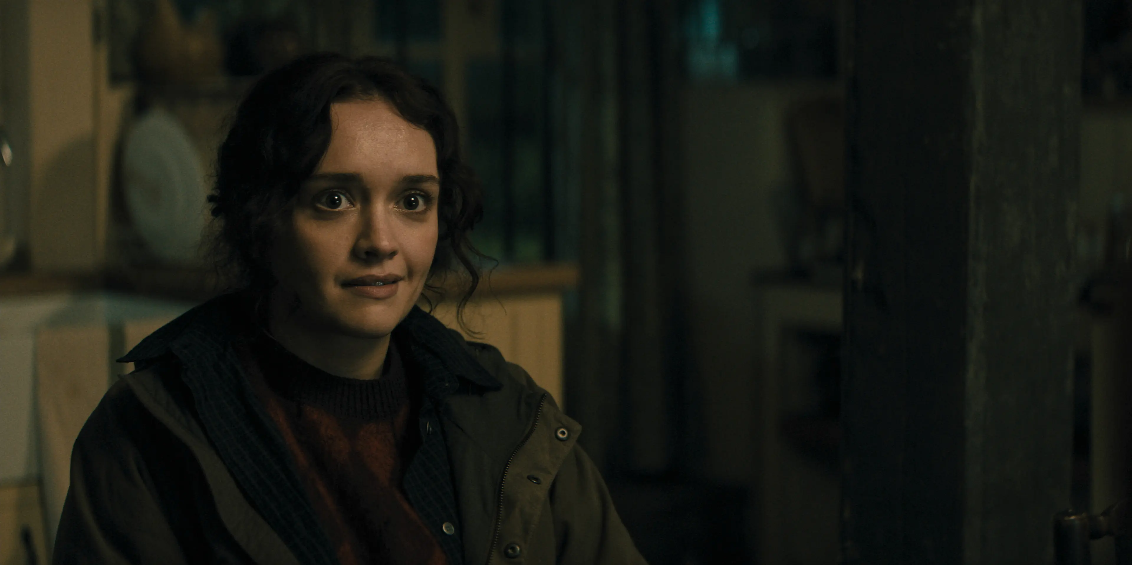 Olivia Cooke as Sid Baker stands in a dim room wearing a dark jacket.