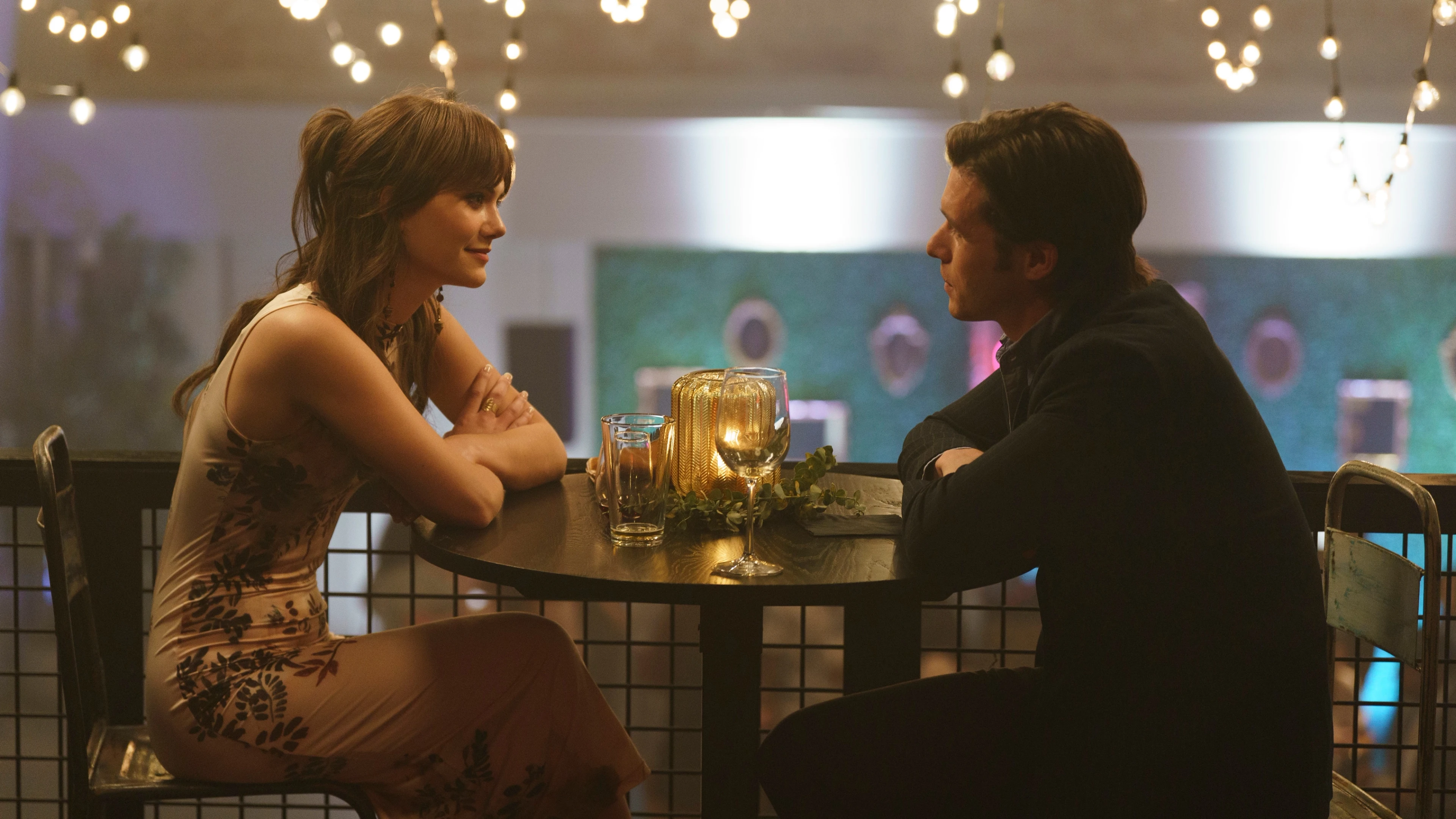 Harper and Charlie sit across from each other at a round table under string lights.