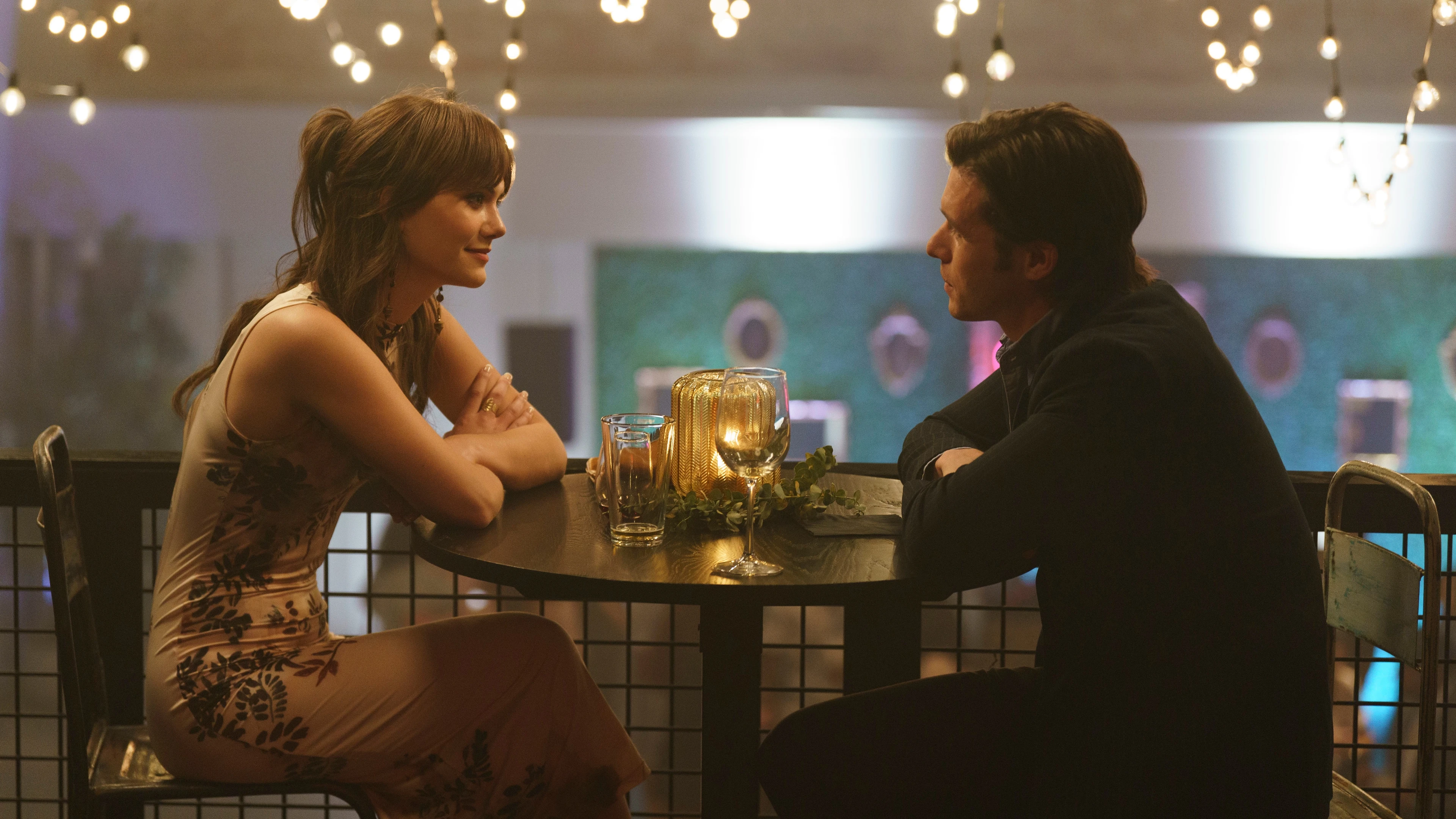 Harper and Charlie sit across from each other at a round table under string lights.