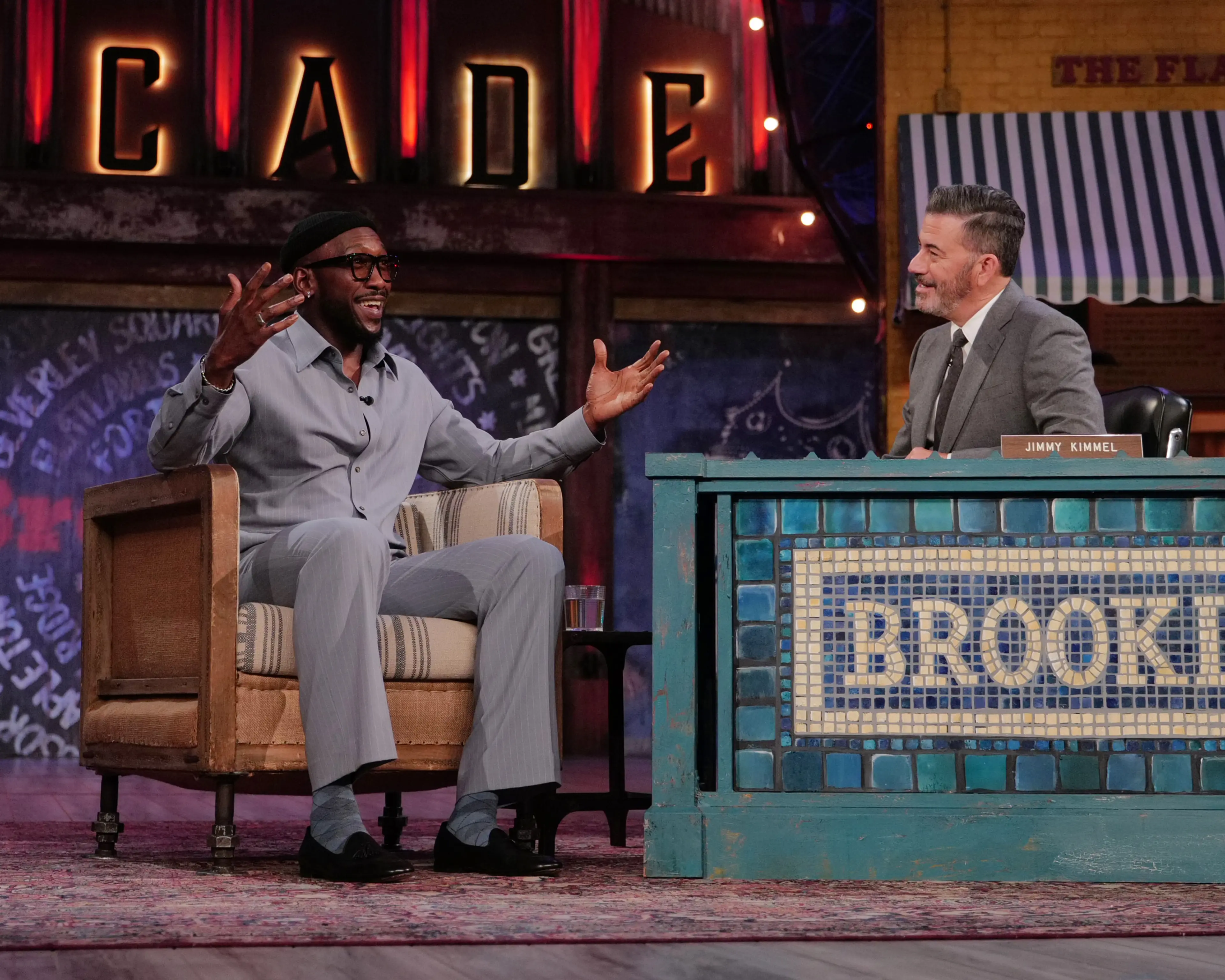 Mahershala Ali gestures with both hands while Jimmy Kimmel smiles at his desk.