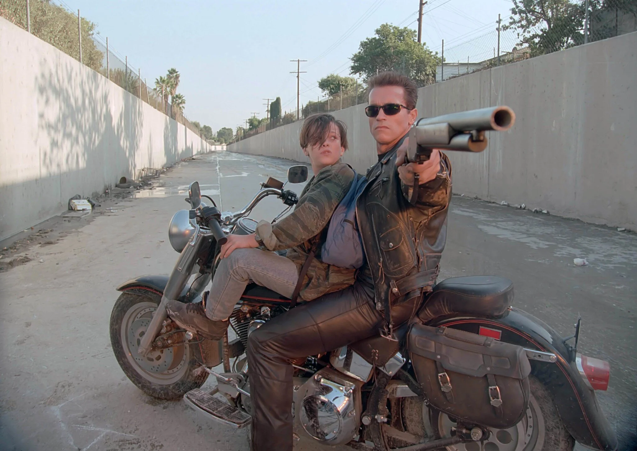 Arnold Schwarzenegger as the T-800 aims a shotgun while John Connor sits on a motorcycle in Terminator 2.