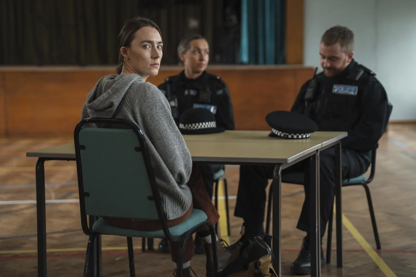 Saoirse Ronan as Maria sits at a classroom table while two officers stand nearby.